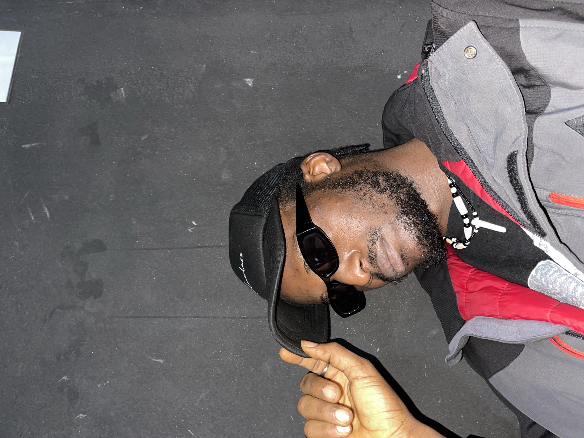 A medium close-up shot shows a Black man wearing a black baseball cap, sunglasses, a black t-shirt, a red and black layered jacket, and a white beaded necklace with a cross pendant. The man is holding the brim of his cap with his left hand. He has a beard and is looking down. The background is a dark, textured wall. The scene is indoors and there is no indication of the city of Jos, Nigeria.