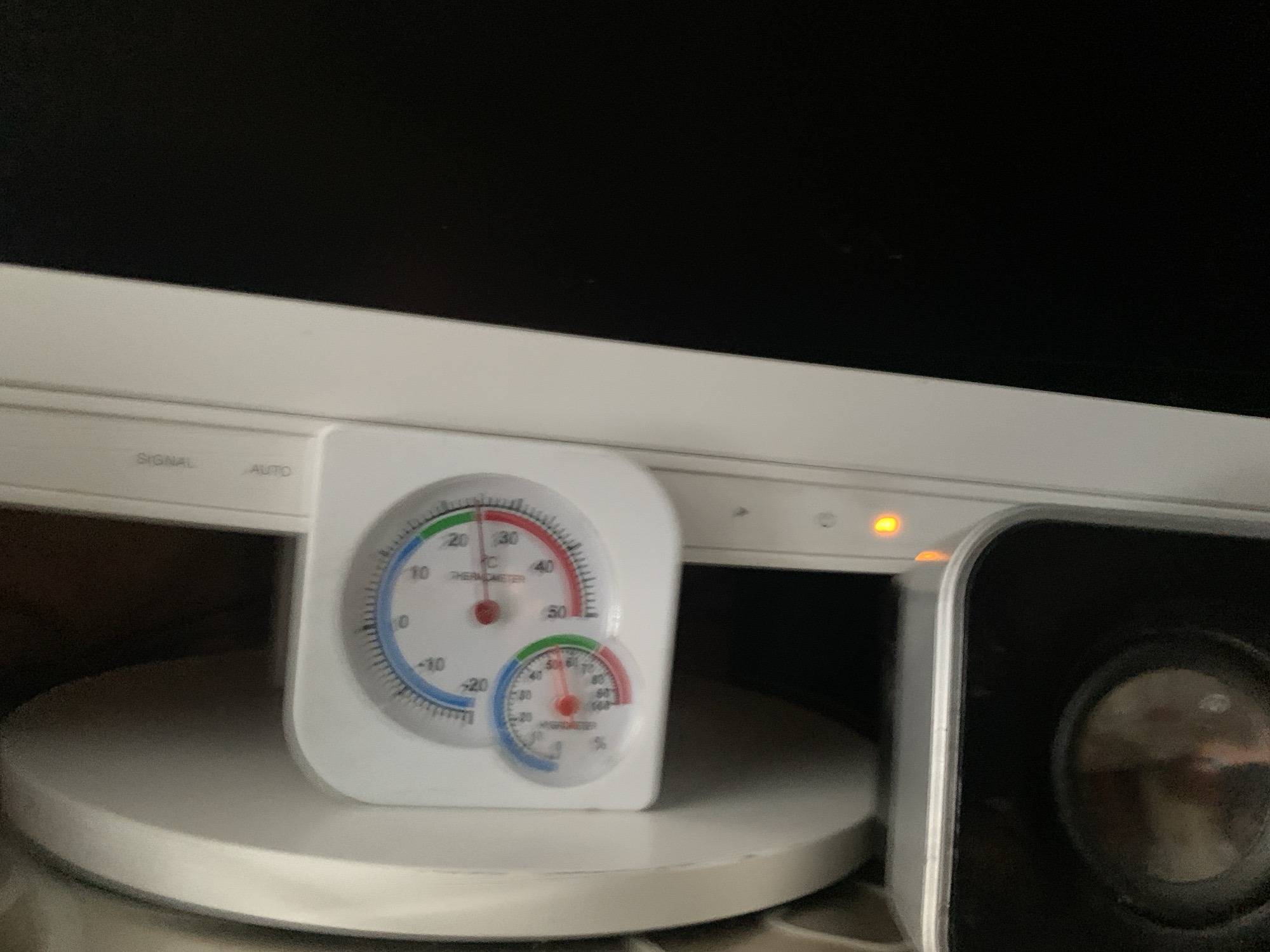 The image shows a close-up of a digital thermometer and hygrometer unit situated on top of a white circular base. The unit has two circular dials, one for temperature and one for humidity. The temperature dial shows a reading of approximately 22 degrees Celsius, indicated by a red needle. The humidity dial shows a reading of approximately 50 percent, also indicated by a red needle. To the right of the thermometer/hygrometer unit, a black speaker with a silver trim is visible. Above the unit, on a white electronic device, two buttons labeled "SIGNAL" and "AUTO" can be seen. An orange indicator light is also present on this device. The background is dark and out of focus. The scene appears to be indoors, likely in a home environment, with no specific indicators pointing to the city of Нижнее Альмурзино, Russia.