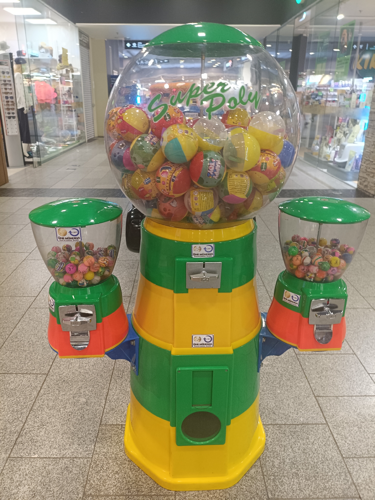 A brightly colored gumball machine stands in a public indoor space, likely a shopping mall in Narva, Estonia. The machine is predominantly yellow and green, with red accents on the side dispensers. The main body of the machine features a large, clear globe filled with colorful bouncy balls, some of which have cartoonish designs. Above the globe, the text "Super Poly" is visible in green lettering. Two smaller transparent dispensers, also filled with bouncy balls and topped with green lids, are attached to the sides of the main unit. Each dispenser has a metal coin slot. The machine is positioned on a light-colored tiled floor. In the background, blurred out, are storefronts and displays, typical of a retail environment. No people are visible in the immediate vicinity of the machine.