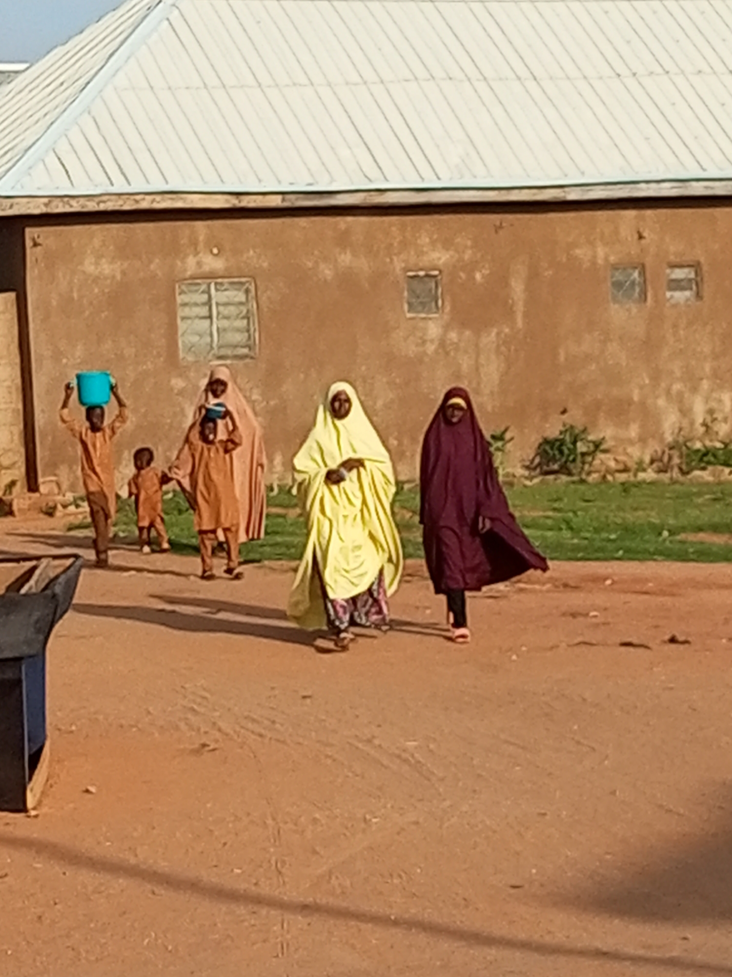In Tempure, Nigeria, several individuals walk along a dirt road in front of a two-story building with a corrugated metal roof. On the left side of the image, a young boy carries a blue bucket on his head, while another young child walks beside him. Behind them, an adult woman in a light pink hijab carries a smaller bucket on her head. To the right of this group, a woman in a bright yellow hijab walks with her head bowed, and further to the right, another woman wearing a dark purple hijab walks with her head covered and a portion of her face obscured, her long garment trailing behind her. The building behind them has several windows with barred openings and appears weathered. The ground is a reddish-brown dirt, and patches of dry grass and some scattered bushes are visible to the right of the building. A dark blue and brown object, possibly a bench or a storage container, is partially visible in the lower left corner. The scene suggests a typical day in a Nigerian community, with people engaged in daily activities.