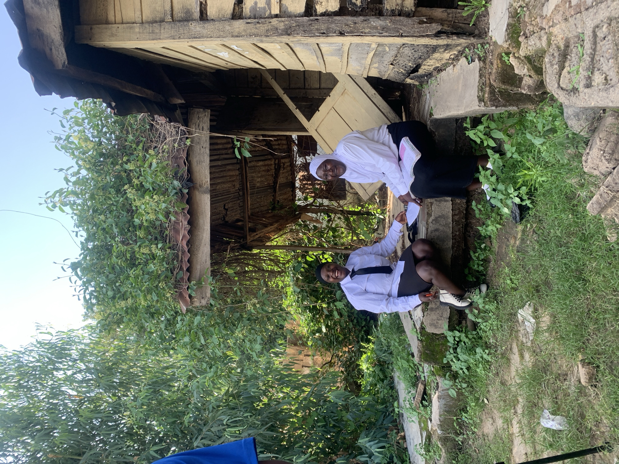 This image, captured in the city of Jos, Nigeria, depicts two adult females seated on a weathered concrete ledge outside a dilapidated wooden structure. The individual on the left, a younger adult, is dressed in a white long-sleeved shirt, a black necktie, a black skirt, and a dark head covering, with white and black boots on her feet. She is smiling broadly and looking towards the viewer, with her left hand gesturing towards a document. The individual on the right, an older adult, wears a white long-sleeved shirt, black trousers or a long skirt, and a white hijab. She is also smiling broadly towards the viewer and holds an open book or document displaying pink highlights. Both women appear to be engaged in an interaction centered around these documents.
The background features a single-story building constructed from weathered, discolored wooden planks, exhibiting signs of age and neglect. Its roof is composed of rusty corrugated metal, heavily overgrown with various green plants and vines. A light-colored wooden door, detached from its frame, is propped diagonally within the main doorway opening of the structure. Dense green vegetation, including tall trees with narrow leaves and numerous shrubs, frames the building and extends into the background. The foreground consists of dirt ground interspersed with patches of grass and weeds, with a piece of discarded white plastic visible on the far left. A partial view of a person in a blue shirt and a black tripod are also present on the far left of the frame. The scene is illuminated by natural daylight under a clear, bright sky.