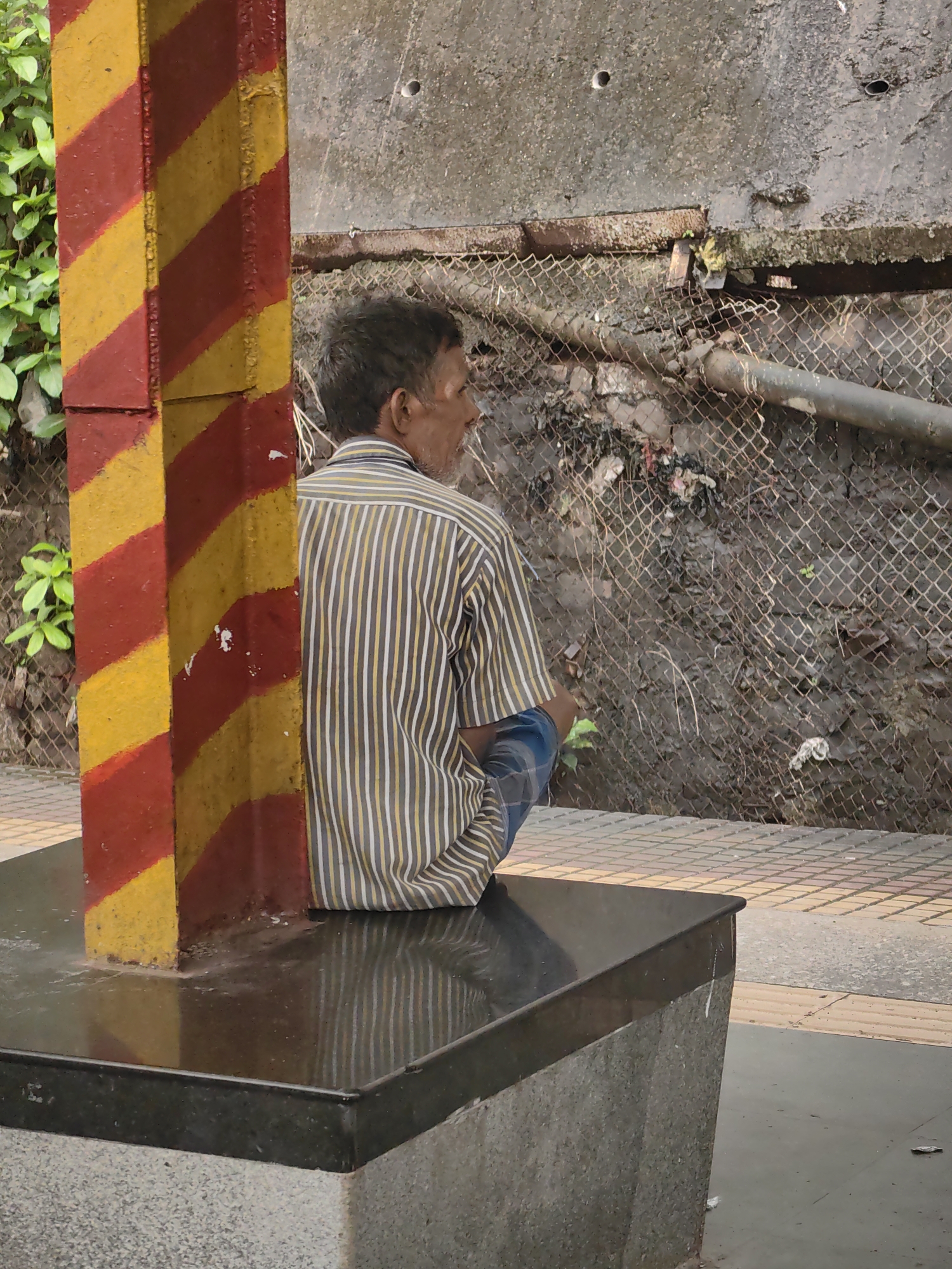 A man is sitting on a black granite bench in Mumbai, India, by a red and yellow striped pillar. He is wearing a striped shirt and blue pants. The background shows a dirty wall with a chain-link fence in front of it. There is debris on the ground behind the fence. The man is looking away from the camera.