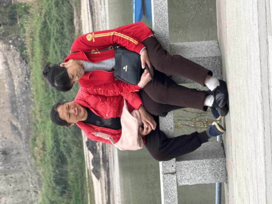 Two East Asian women, appearing middle-aged to elderly, are seated side-by-side on a light gray, rough-hewn stone bench in Shapingba District, China.
The woman on the left wears a red puffer jacket over a black long-sleeved shirt, dark-colored pants, and blue slipper-style shoes with dark socks. A pink shoulder bag is draped over her left shoulder and rests in her lap, where her hands are clasped. Her head is turned slightly to her right, and she exhibits a subtle smile.
The woman on the right wears a red zippered track jacket with yellow/gold stripes on the sleeves and a circular emblem on the left sleeve, over a light gray long-sleeved shirt, dark brown pants, and black closed-toe shoes with white socks. A black shoulder bag with a light-colored emblem rests on her lap. She holds a dark-colored smartphone or similar device with both hands, looking downwards at it. Her hair is pulled back, forming a small bun.
The immediate foreground consists of a light-colored paved surface. In the mid-ground, a calm, greenish-grey body of water is visible. The background features a distant bank with green foliage, patches of dark exposed earth or debris, and a light-colored concrete retaining wall. A small blue object, possibly part of a boat, is partially visible on the water to the far right.