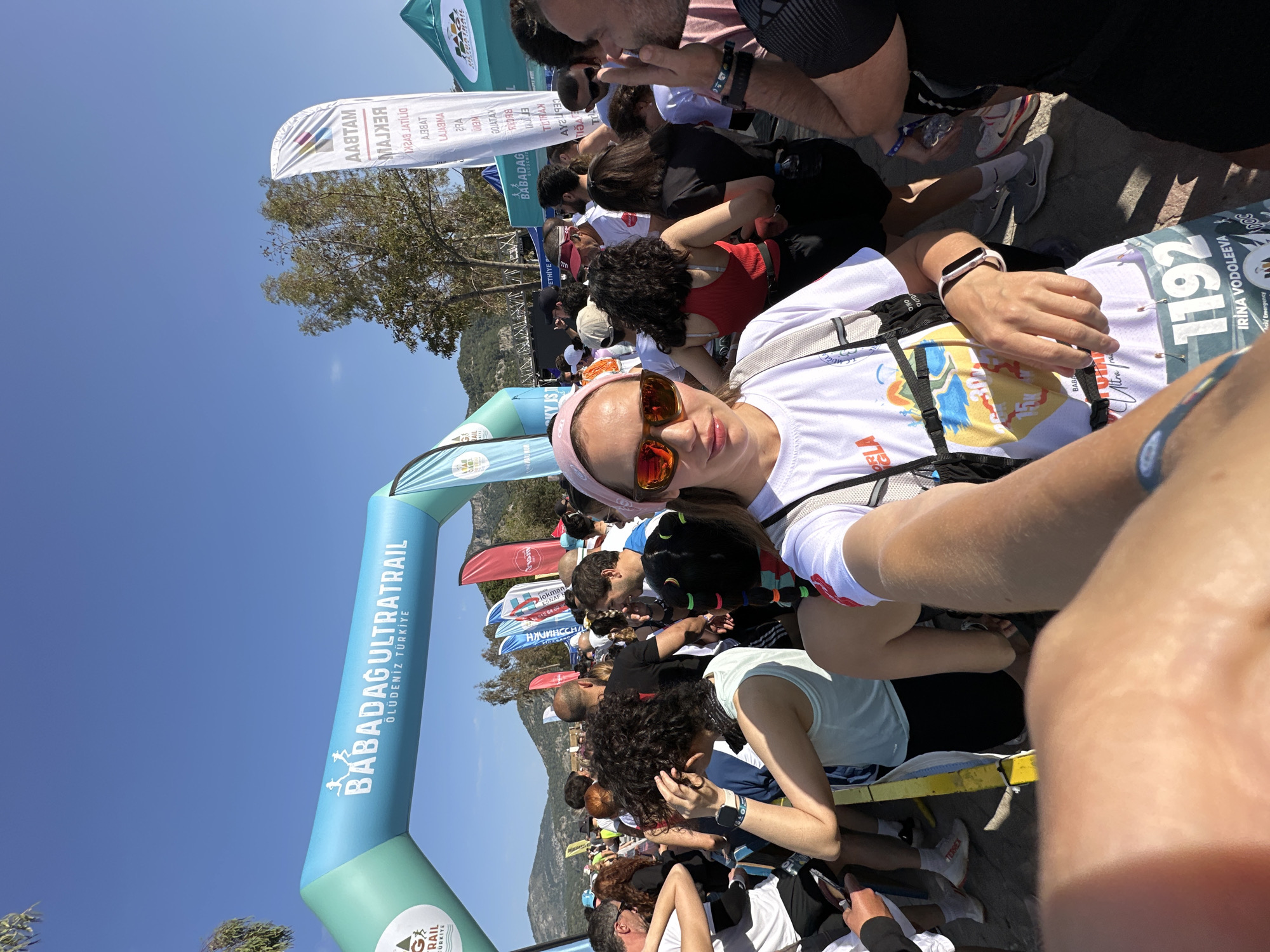 Here is a detailed description of the image:
**Scene:** The image appears to be taken at the "BABADAG ULTRATRAIL" event in Ölüdeniz, Fethiye, Turkey. This is indicated by the text on the inflatable archway in the background.
**People:** The image shows a group of people gathered, including the person taking the photo. The selfie focuses on a woman wearing a pink headband, sunglasses, and a white t-shirt with a race number "1192" indicating that she is likely a participant in the ultra trail. Other individuals can be seen behind her, some of whom are seated.
**Objects:** Other objects include race flags, trees, and some sort of barrier or seating that the people are using.
**Action/Interaction:** The primary action visible is the woman taking a selfie. The other people in the frame appear to be resting or preparing for the ultra trail.
**Environment:** The environment is sunny and outdoors.