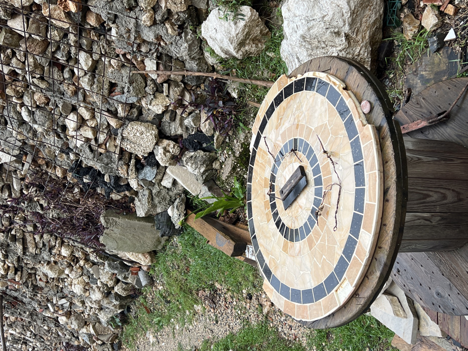 The scene depicts an outdoor area in Rafailovići, Montenegro. In the foreground, a large, weathered wooden cable spool serves as a table base. Resting atop the spool is a circular mosaic tabletop composed of tan/beige and black tiles arranged in concentric circles. The mosaic shows signs of wear, with several tiles missing along its outer edge. Centrally positioned on the mosaic are two small, dark, rectangular wooden blocks, possibly joined, accompanied by several thin, twisted strands of dark, rusty wire. A small, smooth, pinkish-brown pebble is visible on the mosaic's edge.
The background features a steep, man-made retaining wall constructed from an assortment of irregularly shaped light-colored stones, concrete fragments, and darker debris, some held in place by sections of metal mesh or rebar. Sparse patches of dark reddish-purple leafy plants and lighter green foliage grow amidst the stones and along the base of the wall. The ground immediately surrounding the table and extending towards the left is covered with patchy green grass, soil, and scattered small stones, forming a narrow pathway. Various pieces of debris, including a short wooden beam and additional stones, are visible on the ground. A large, light-colored boulder is present on the right side of the image, near the table.
