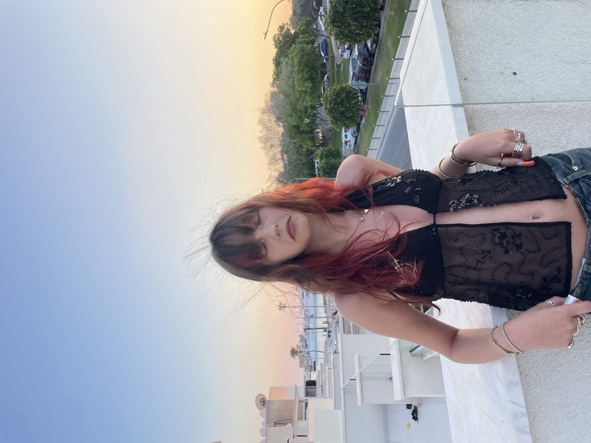 A young woman stands on a rooftop balcony in Paphos Municipality, Cyprus, at sunset. She has long, wavy, dark hair with streaks of red and bangs. She is wearing a black, sheer, sequined halter top and denim jeans. Her eyes are closed, and her head is tilted slightly down. The background shows modern white buildings, palm trees, a street with parked cars, and a body of water with boats in the distance. The sky is a gradient of blue and orange, typical of sunset. The woman is leaning on the balcony railing, which is made of marble and glass.