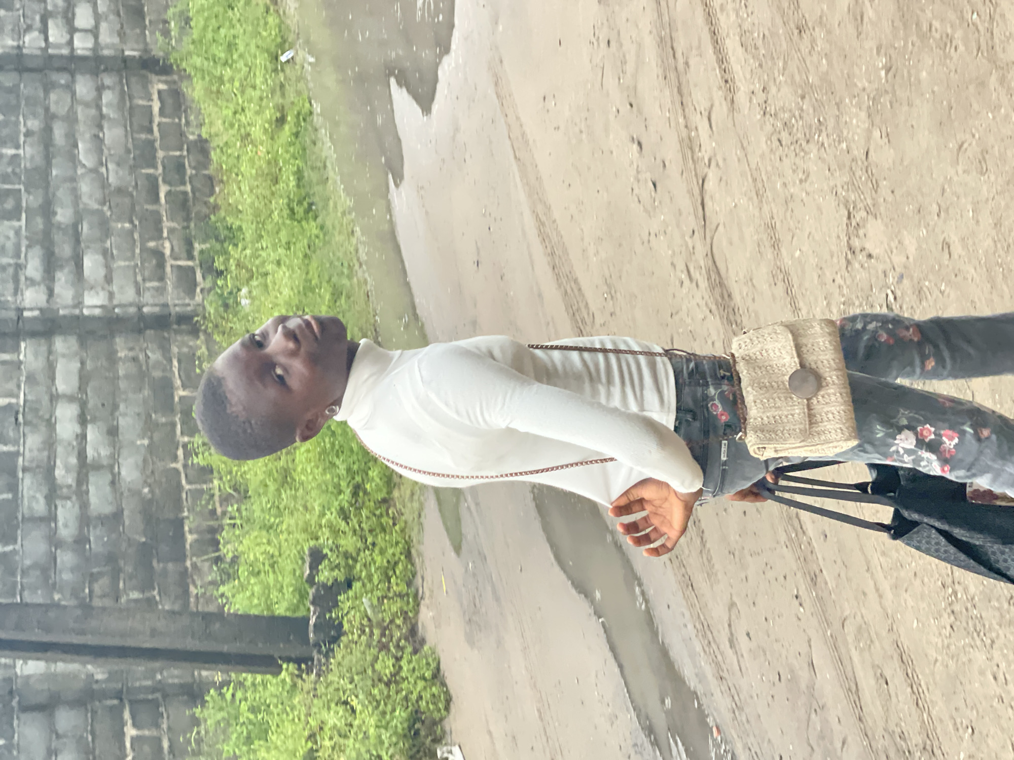 A young person stands on a wet, dirt road in what appears to be a residential area. The person is wearing a white turtleneck shirt, floral print pants, and a small woven purse over their shoulder. They are also carrying a large bag. To the left of the road is a grey brick wall and some green foliage. The ground appears to be muddy, with puddles of water. The scene suggests a recent rainfall. The specific location in Badagry, Nigeria cannot be determined with only this image.