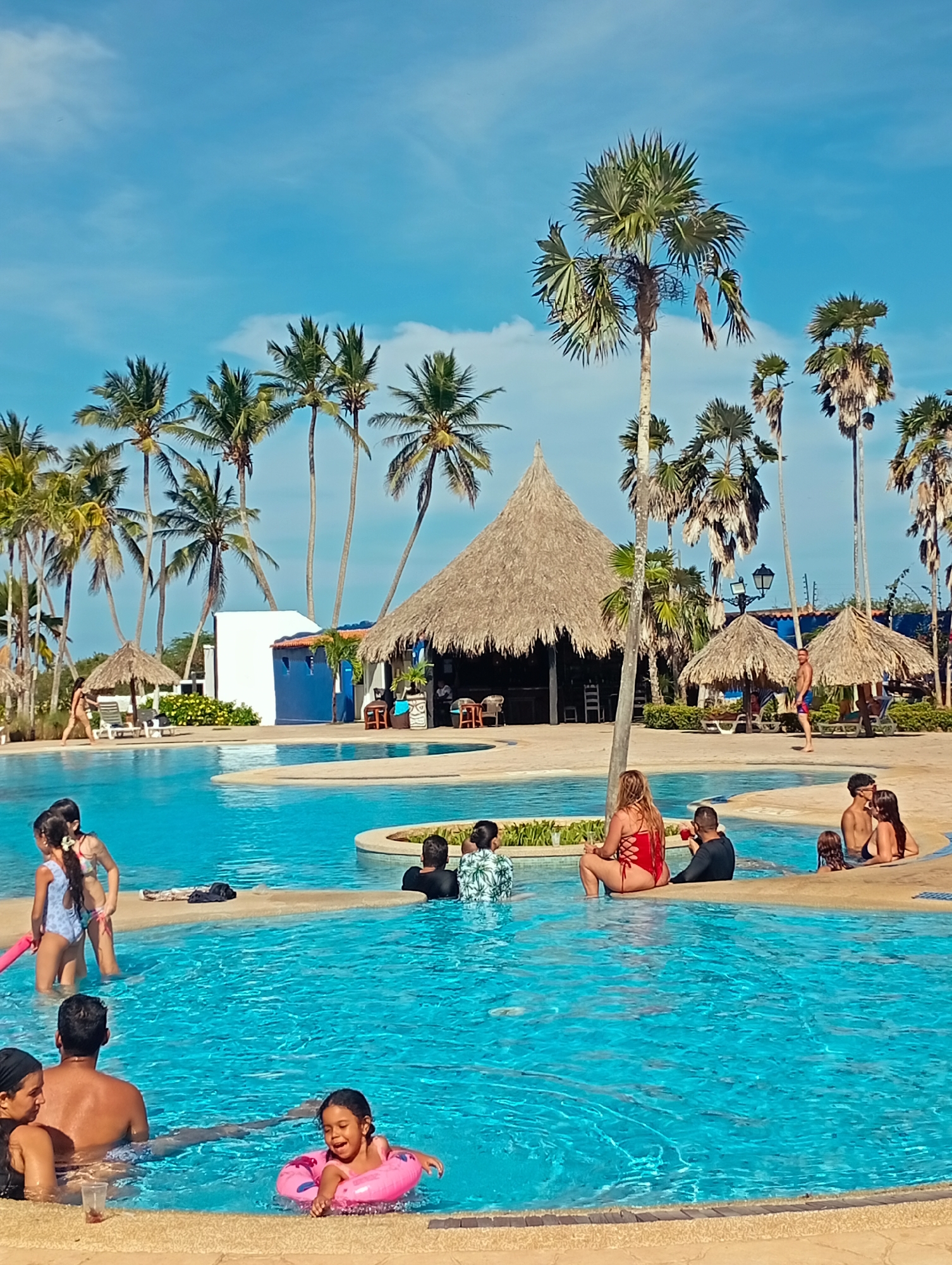 This vibrant image depicts a bustling resort swimming pool complex under a clear blue sky, characteristic of a tropical destination like Playa El Agua, Venezuela. The large, irregularly shaped pool is filled with clear, turquoise water, and is surrounded by numerous tall palm trees that provide a lush backdrop. Several thatched-roof structures, known as churuatas or palapas, are visible; a prominent one in the mid-ground appears to be a bar or restaurant, while smaller ones serve as shaded areas.
Numerous people, including adults and children, are enjoying the facilities. In the foreground and mid-ground, adults are seen relaxing in the water or sitting at the pool's edge, some engaged in conversation. A young girl, smiling, floats in a pink inflatable ring in the foreground. Other children are playing in the water, including two girls near the left side. A woman in a bright red swimsuit sits at the pool's edge on the right, near a man. Further back, a couple is embracing in the water. In the background, people are walking near the thatched huts and sun loungers.
The time of day is clearly daytime, with bright sunlight creating distinct shadows, and the weather is sunny with a few wispy clouds in the expansive blue sky. The general mood conveyed is one of leisure, happiness, and relaxation. There is no discernible text visible in the image.