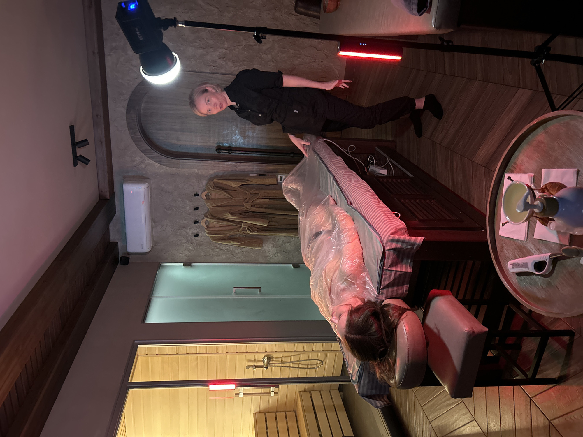 The image depicts an indoor scene in what appears to be a spa or wellness center in Izhevsk, Russia. A woman in black workwear stands overseeing another woman lying on a bed draped with clear plastic. The woman on the bed is positioned near what appears to be a sauna, visible through a glass door. Above, a collection of brown robes hang on hooks. The lighting is artificial, with a studio light on a stand positioned near the standing woman and a red light panel visible in the background. A round table with a tea set is positioned to the right of the bed.