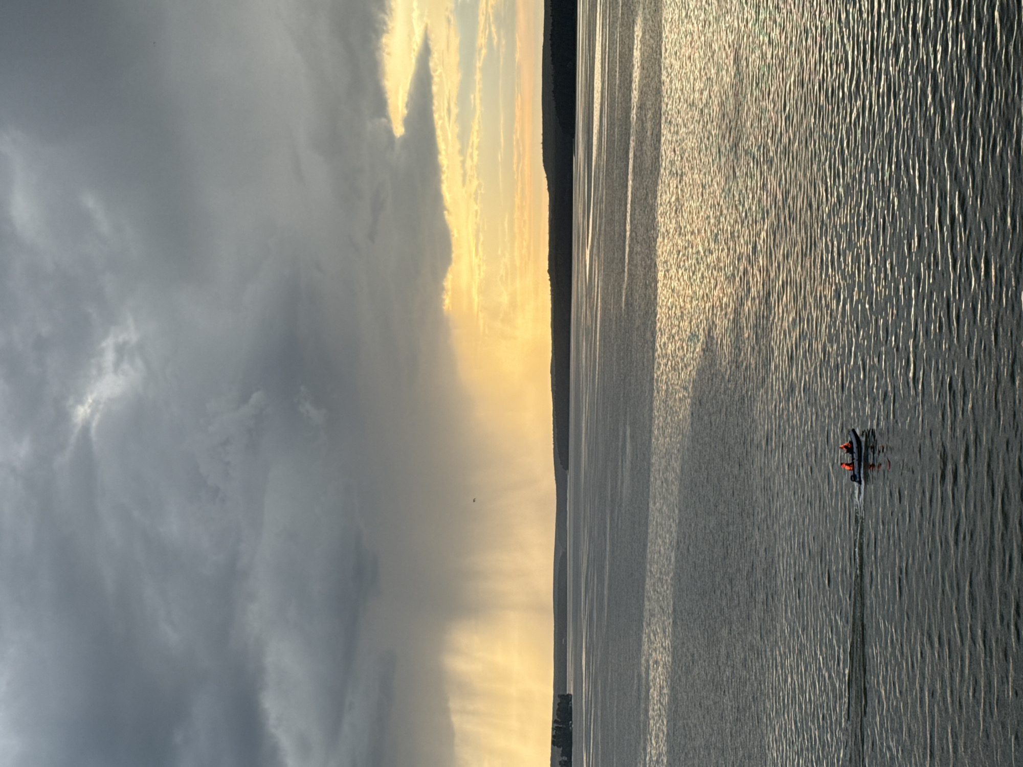 This media file depicts a serene scene on a body of water, likely a lake or river, in Votkinsk, Russia. The composition is split, with a dramatic sky dominating the left side and the calm water filling the right.
The sky is a mix of dark, heavy clouds on the left, suggesting impending rain or a recent storm, transitioning to brighter, golden hues of sunset or sunrise on the right. Sunbeams break through the clouds, casting a warm glow across the water's surface. The water itself is textured with ripples, reflecting the light and creating a shimmering effect. A dark, silhouetted shoreline runs along the horizon, adding depth to the scene.
In the water, a small boat with two individuals wearing orange life vests is visible. They appear to be moving, leaving a subtle wake behind them, suggesting they are engaged in an activity like fishing or leisurely boating.
The time of day is clearly either early morning or late evening, indicated by the dramatic interplay of light and shadow in the sky. The weather appears to be changing, with the possibility of rain from the darker clouds juxtaposed with the warmth of the setting or rising sun.
No visible text is present in the image. The overall mood is tranquil and evocative, capturing the beauty of nature at the edge of the day.
