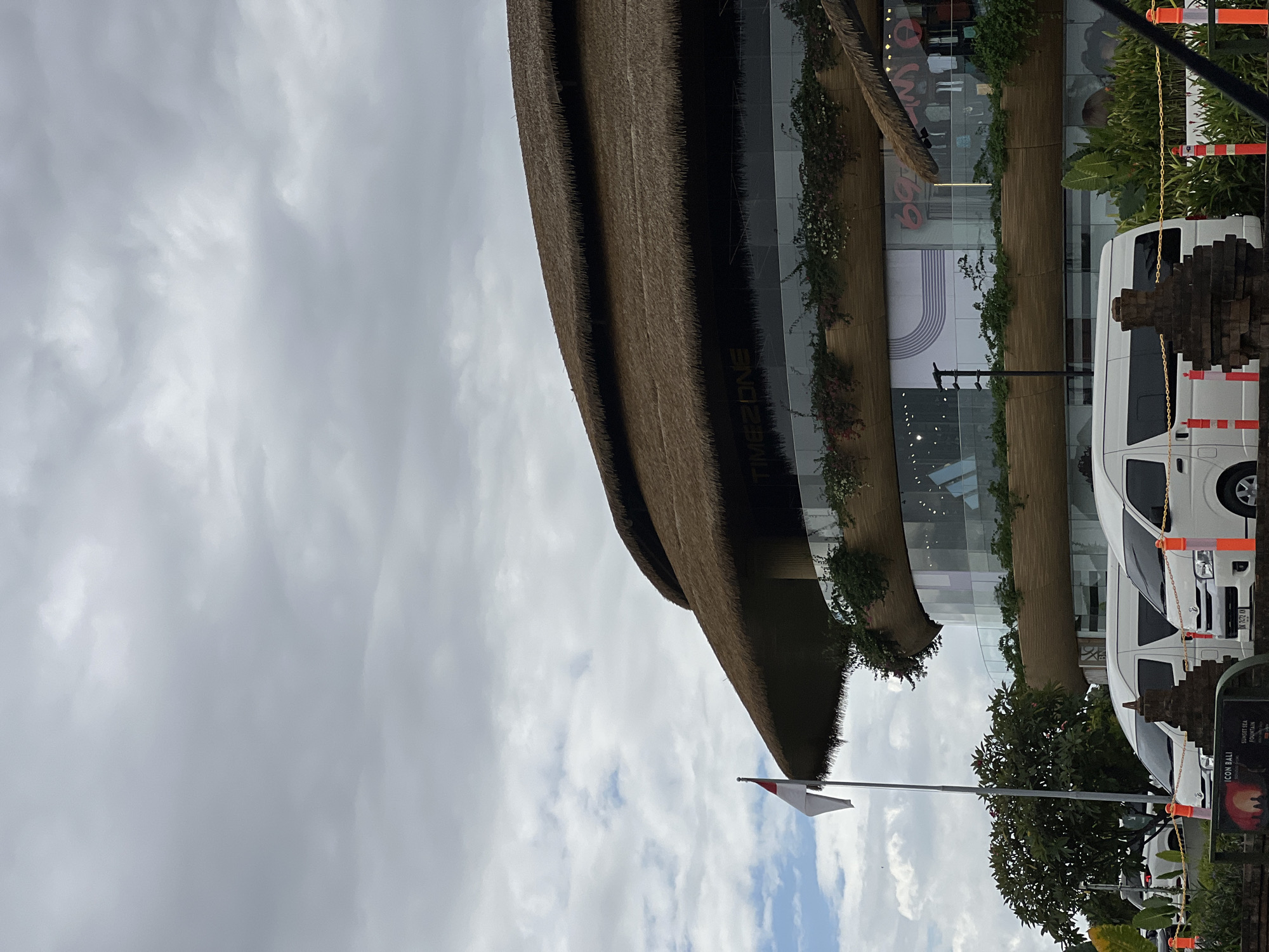 The image shows the exterior of a modern building with a thatched roof in Denpasar, Indonesia. The building features a curved facade with multiple levels of glass windows, some of which display advertisements. The thatched roof is a prominent architectural element, giving the building a distinct tropical aesthetic.
In front of the building, there are several white vans parked, suggesting a transportation hub or a commercial establishment catering to visitors. A flagpole with the Indonesian flag, red and white, stands to the left of the building, indicating its location in Indonesia. The sky is overcast, with a mix of gray clouds and patches of lighter sky, suggesting a partly cloudy day. Greenery, including trees and plants, surrounds the building and parking area, adding to the natural setting.
The overall scene depicts a commercial or entertainment complex with a unique architectural design that blends modern and traditional elements, set against a tropical backdrop under a cloudy sky.