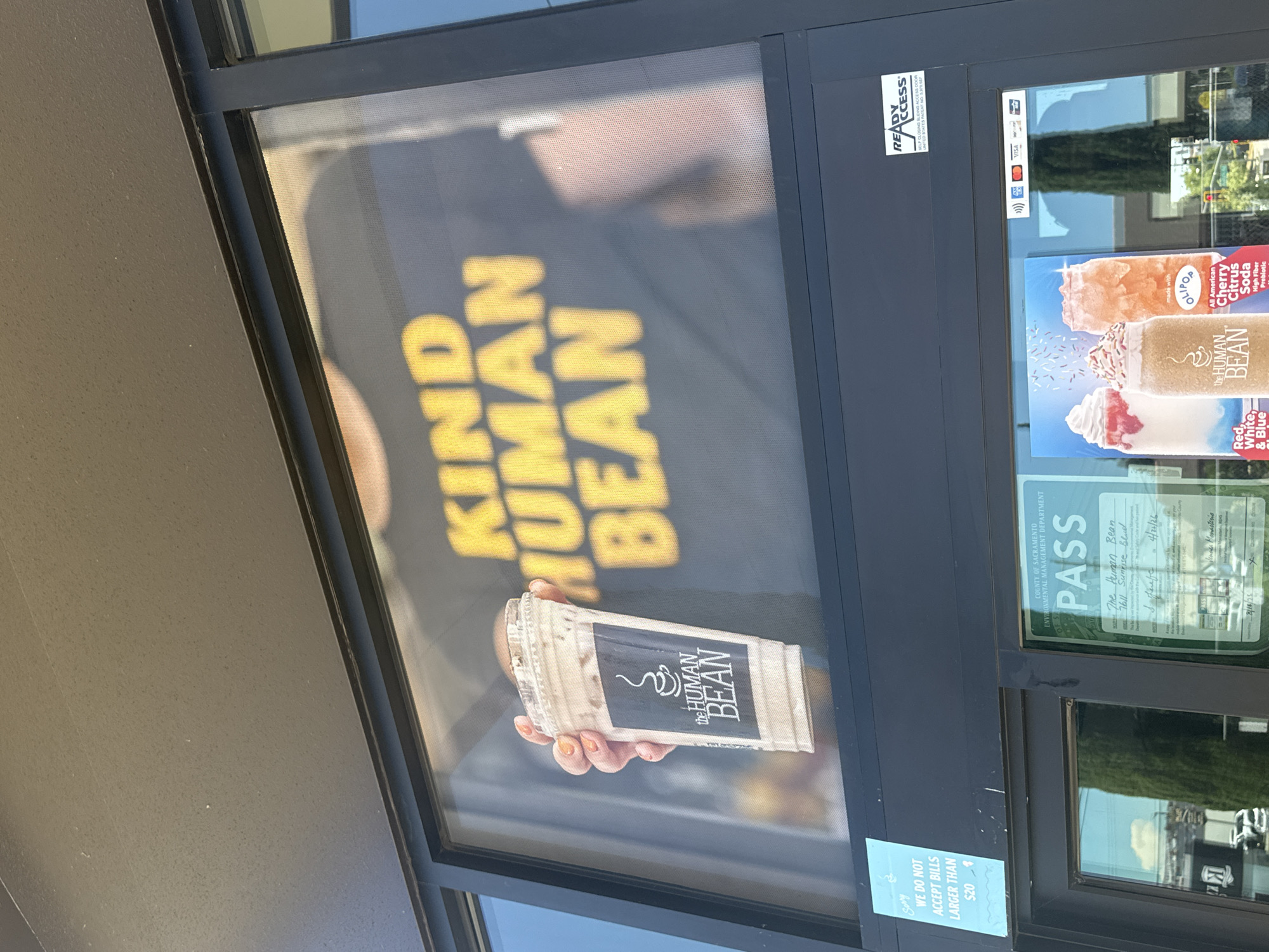 A medium shot captures a drive-thru window at "The Human Bean" coffee shop, located at 7611 Sunrise Blvd, Citrus Heights, United States of America. The window displays a large graphic of a hand holding a cold beverage with the "The Human Bean" logo. Behind the window, a person wearing a black t-shirt with the words "KIND HUMAN BEAN" in large yellow letters is visible. A "PASS" sticker from the County of Sacramento Environmental Management Department is affixed to the lower right portion of the window, dated 4/23/20. A smaller blue sign to the left states, "Sorry, WE DO NOT ACCEPT BILLS LARGER THAN $20". A "READY ACCESS" sticker is on the upper right of the drive-thru unit. Below the window, promotional images of iced beverages are displayed.