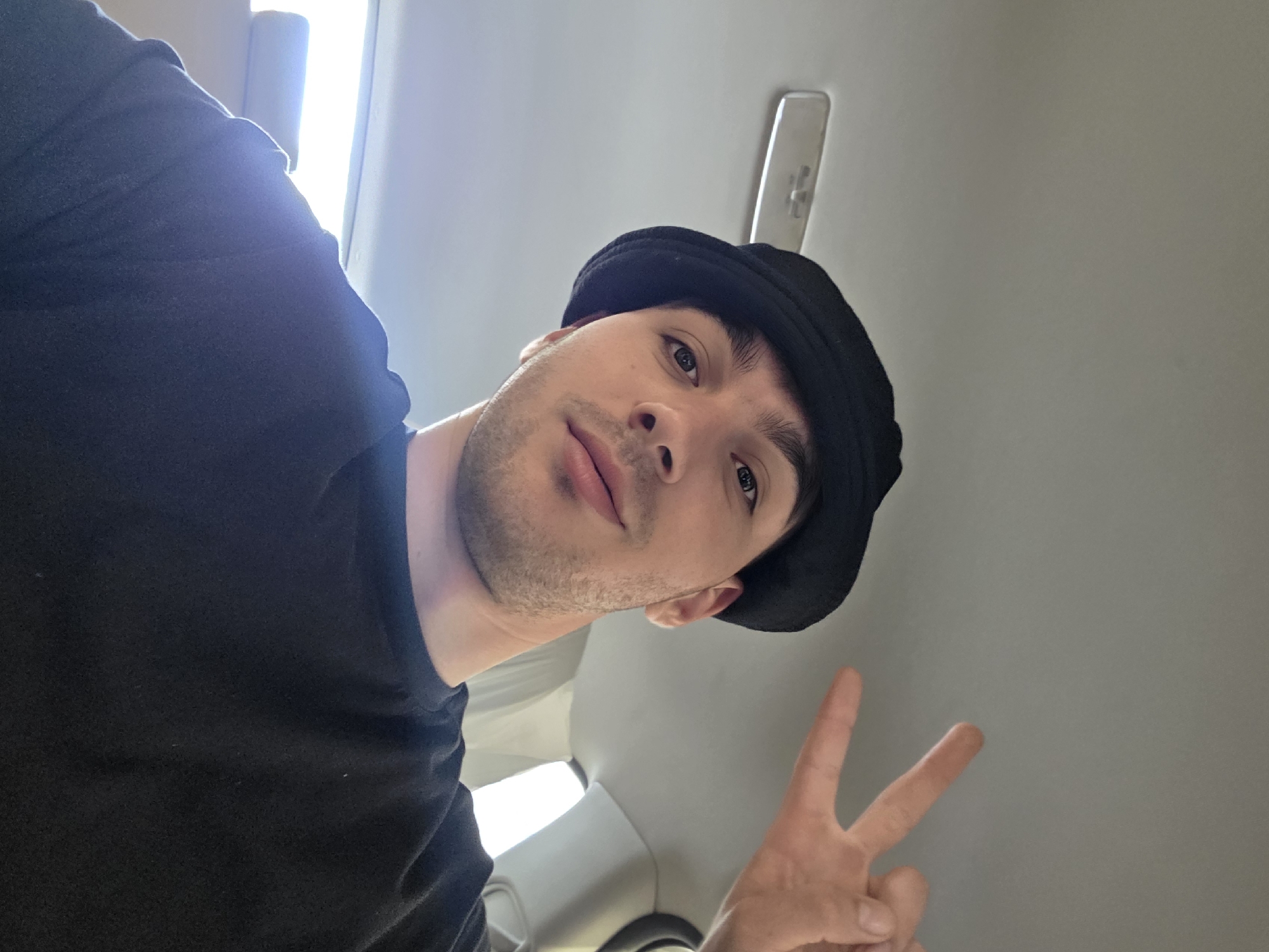 A man wearing a black bucket hat and a black t-shirt is captured in a selfie from the chest up. He has a light complexion, dark hair, and is looking towards the camera with a slight smile. His right hand is raised, forming a peace sign. The background appears to be the interior of a vehicle, with a grey headliner and a light fixture visible. The lighting suggests it may be daytime.