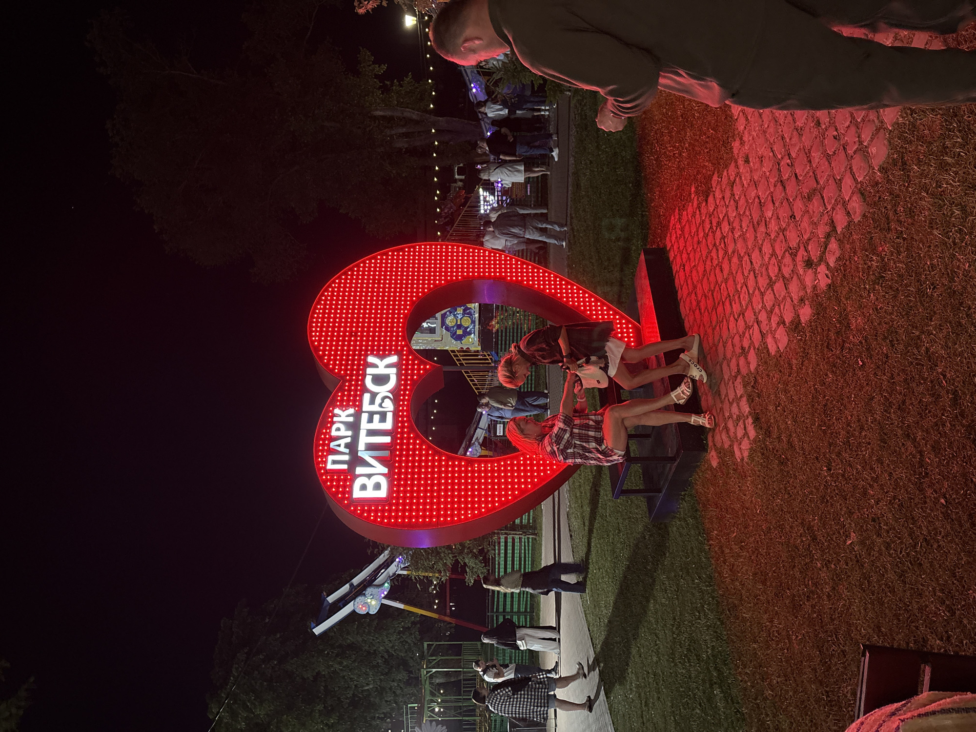The image depicts an outdoor nighttime scene in Vitebsk Park, Vitebsk, Belarus. A large, illuminated red heart-shaped sign with white lettering reads "ПАРК ВИТЕБСК" (Park Vitebsk). In the foreground, two young women are seated on a dark bench in front of the heart. The woman on the left, wearing a plaid shirt and shorts, is looking down. The woman on the right, in a darker top and shorts, is reaching into a white bag. To the right of them, a man in a dark sweatshirt and pants stands with his back to the camera, looking at something in his hands.
In the background, other park-goers are visible. To the left, a swing ride is partially visible, illuminated by colorful lights. Further back, people are standing and walking near amusement park attractions. The ground is a mix of grass and cobblestone paving, illuminated by the red glow of the heart sign and string lights in the background. Trees are scattered throughout the park, silhouetted against the night sky.