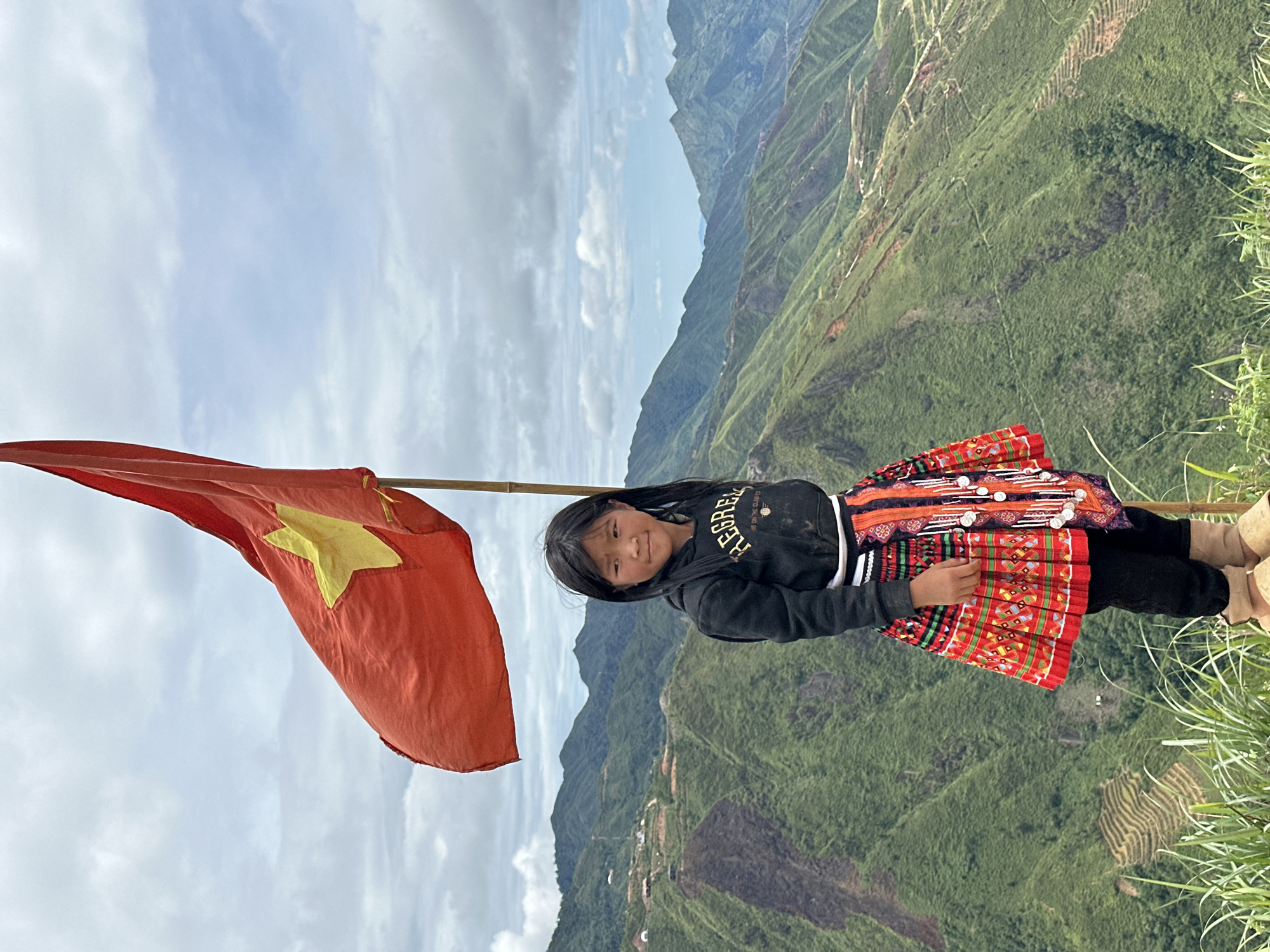 A young girl stands on a grassy overlook in Ta Xua, Vietnam, with the Vietnamese flag flying behind her. She is wearing a black hooded sweatshirt and a colorful, patterned skirt. The landscape behind her is mountainous and green, with terraced fields visible on the slopes. The sky is cloudy.