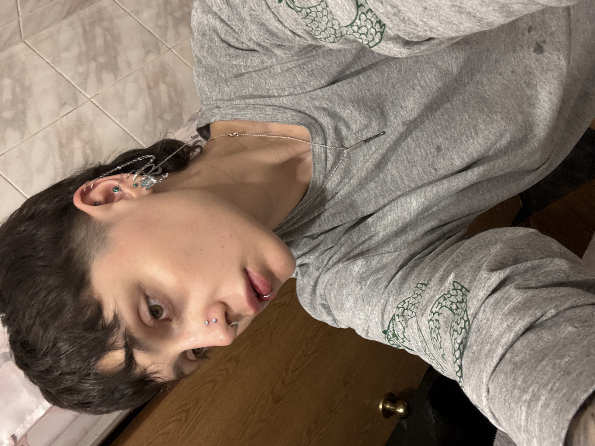 A person with multiple piercings is captured in a photograph. The piercings are visible on the ear, nose, and lip. The person is wearing a gray long-sleeved shirt with a design, possibly a stylized image of a hop plant, on the sleeves. The background consists of tiled flooring and a wooden door. There is no information in the image to indicate that the location is in the city of Saint Petersburg, Russia.