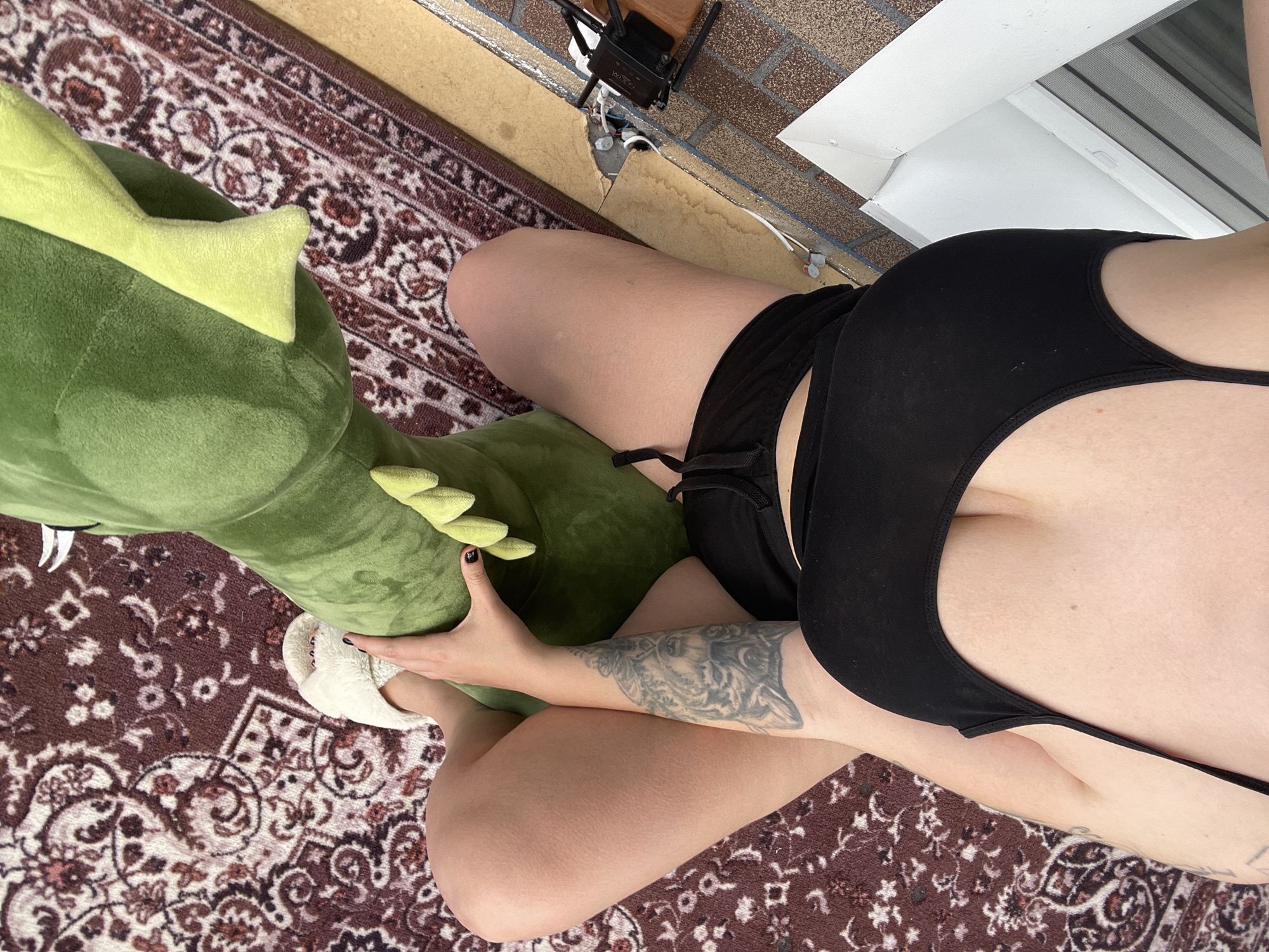 The image shows an overhead view of a person seated on a patterned rug, possibly outdoors near a brick wall and a white window frame. The person is wearing black attire. The location appears to be in Мосметаллоконструкция Онт, Russia. The individual is holding a large, green, plush dragon toy, with their feet clad in white slippers. A router is visible near the brick wall.