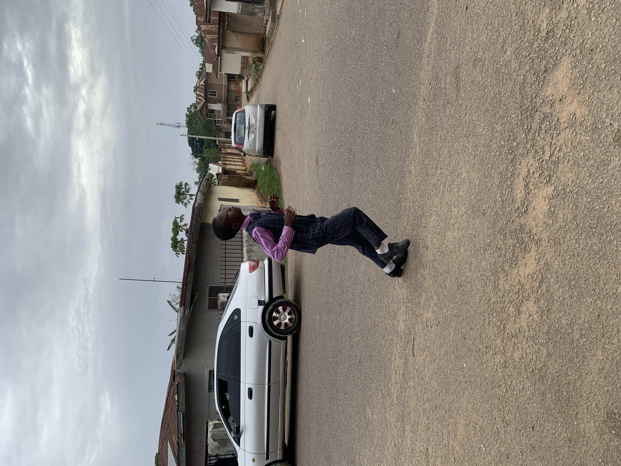 A man is pictured on a street in Jos, Nigeria. He is wearing a pink shirt, a dark pinstripe vest, and matching pants. He is also wearing black shoes with white socks. He appears to be frozen mid-motion.
The background consists of a paved street, various buildings, and two parked cars: a white sedan and a silver SUV. A utility pole with overhead wires is visible. The sky is overcast. The buildings suggest a residential or mixed-use neighborhood.
