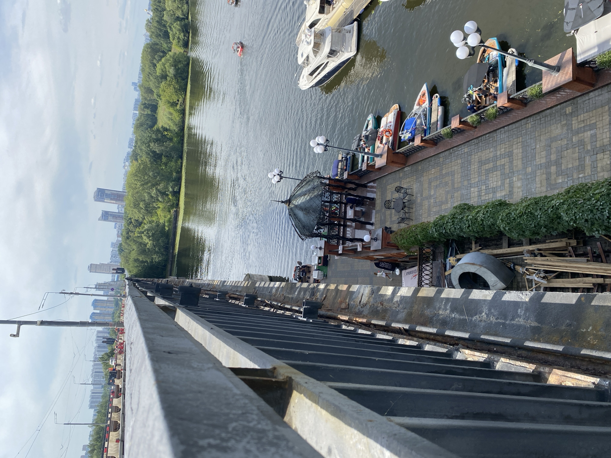 The image shows a view from a bridge looking down at a waterfront area. A paved walkway lines the edge of the water, with several boats docked along it. Among the boats are speedboats, pedal boats, and a larger yacht. There is a gazebo with tables and chairs on the walkway, suggesting it's a recreational or dining area. A few people are visible on the walkway and on some of the boats. Beyond the waterfront, there is a line of trees, and in the distance, city buildings can be seen. The sky is cloudy. The bridge itself has a metal railing and a concrete structure. Construction materials, including wooden planks and a large concrete ring, are piled on the side of the walkway near the bridge.