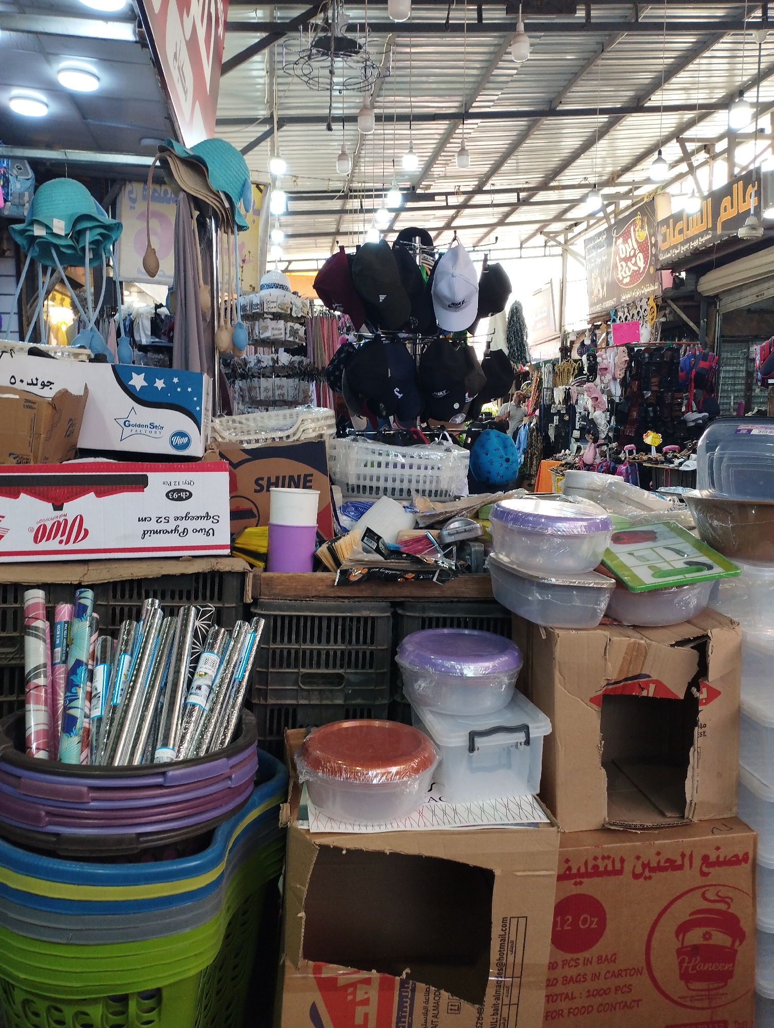 The image depicts a market stall in Baqa'a camp, Jordan, overflowing with merchandise. Various goods are displayed, including an assortment of hats, plastic storage containers of different sizes and colors, and what appear to be cleaning supplies like mops. Cardboard boxes, some open and revealing their contents, are stacked around the stall. In the background, more items are visible on shelves and racks, including clothing and possibly accessories. The stall appears to be covered by a corrugated metal roof, with exposed ceiling structures and lighting fixtures. The overall impression is one of a busy, densely packed retail environment characteristic of a local market.