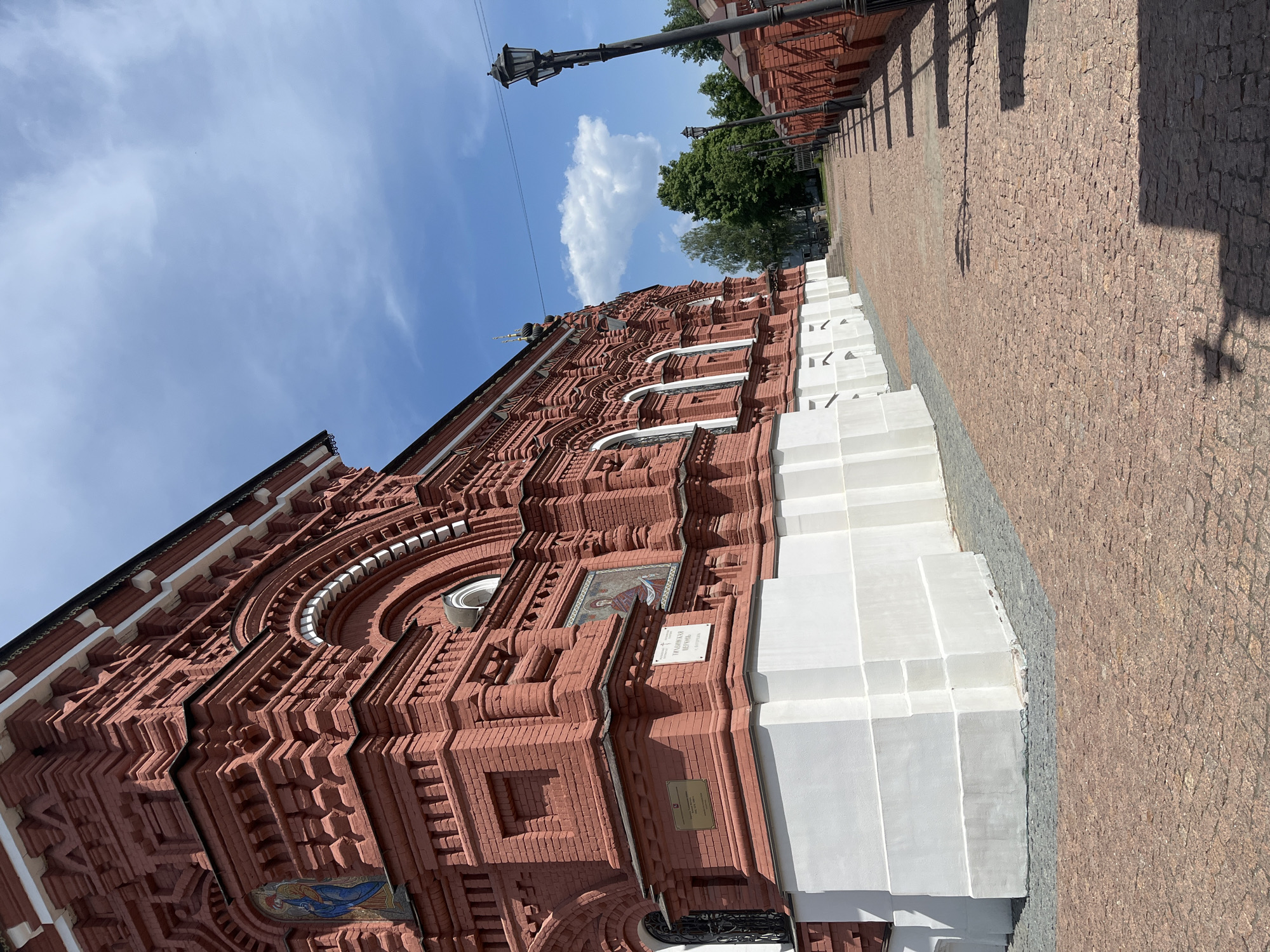 The image shows the façade of a brick building, possibly a church, with ornate red brickwork and white accents. The building has arched windows, mosaics, and plaques affixed to its exterior. The façade is set against a bright blue sky with a single, puffy white cloud. A cobblestone walkway stretches along the side of the building, and in the distance, streetlights and trees are visible. The location is Unknown, Unknown.