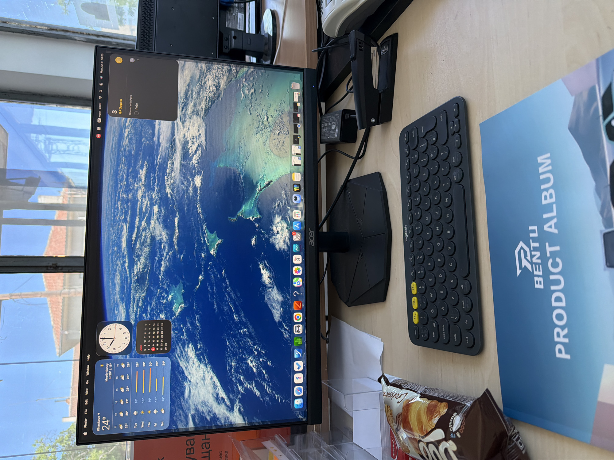 An indoor, eye-level, medium shot shows a computer monitor displaying a desktop with widgets for weather, clock, and calendar. The monitor sits on a wooden desk in front of a window that offers a view of a Mediterranean-style building with red-tiled roofs. To the left of the monitor, on the desk, is a bag of croissants. To the right, a black stapler sits next to a stack of papers. A black Logitech keyboard is positioned in front of the monitor, and a blue and white brochure titled "BENTU PRODUCT ALBUM" is placed in the foreground. The monitor displays a desktop background of Earth from space. The screen's dock shows various application icons including Finder, Safari, Mail, Calendar, Photos, and a trash can. The system status bar displays the time as Monday, June 8, 10:00 AM.