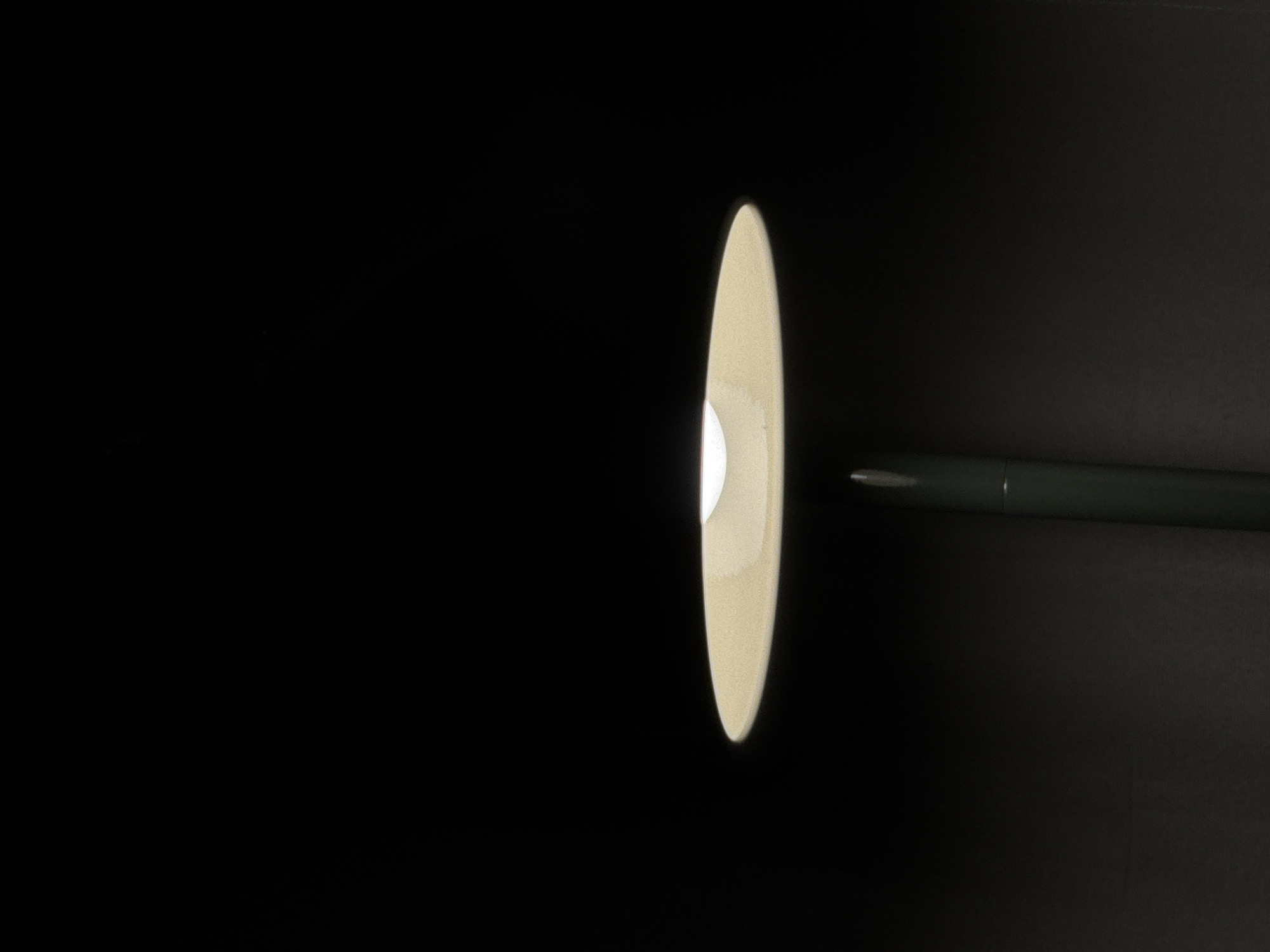 In the darkness of an unknown location, a single lamp emits a soft, warm light from its circular shade. The shade is a pale yellow or cream color, and its underside is illuminated by a bright white light source, likely an LED bulb, which is partially visible. The lamp has a slender, dark green stem that extends downwards out of frame. The background is completely black, suggesting a very dimly lit or completely dark environment. There are no other objects or people visible in the scene.