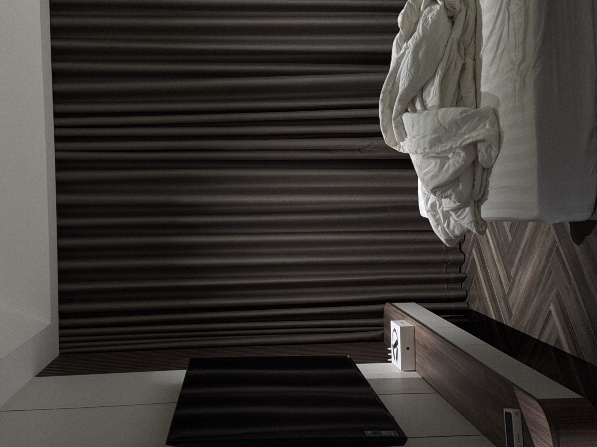 The image depicts an interior scene. The dominant feature is a bed, which is partially visible, with white bedding and a crumpled white duvet. Adjacent to the bed is a wooden headboard or side table with a white surface. A box, possibly packaging for headphones, is placed on the headboard, with the headphone image visible on the box. A black rectangular object, likely a television screen, lies on the floor in front of the headboard. The background is a wall with a textured, horizontal stripe pattern. The overall color palette is muted, with shades of gray, white, and brown. The location and city are not discernible from the image.