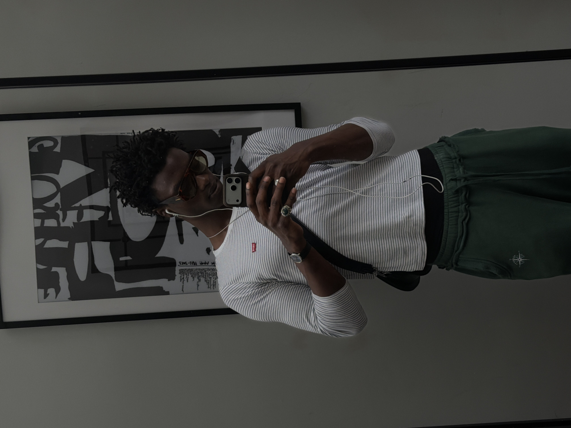A dark-skinned man is taking a selfie in front of a gray wall, presumably indoors. He is wearing a striped, long-sleeved shirt with a small red tag on the chest, green sweatpants with a logo on the left thigh, and headphones with a wire running down his chest. He also has sunglasses and a watch on his wrist, along with rings on his fingers. Behind him is a framed black and white abstract print on the wall, and there's a black horizontal stripe near the top of the frame. The location appears to be indoors and judging from the clothing, the subject is in London, United Kingdom, where it is trendy to wear sweatpants or jogging bottoms.