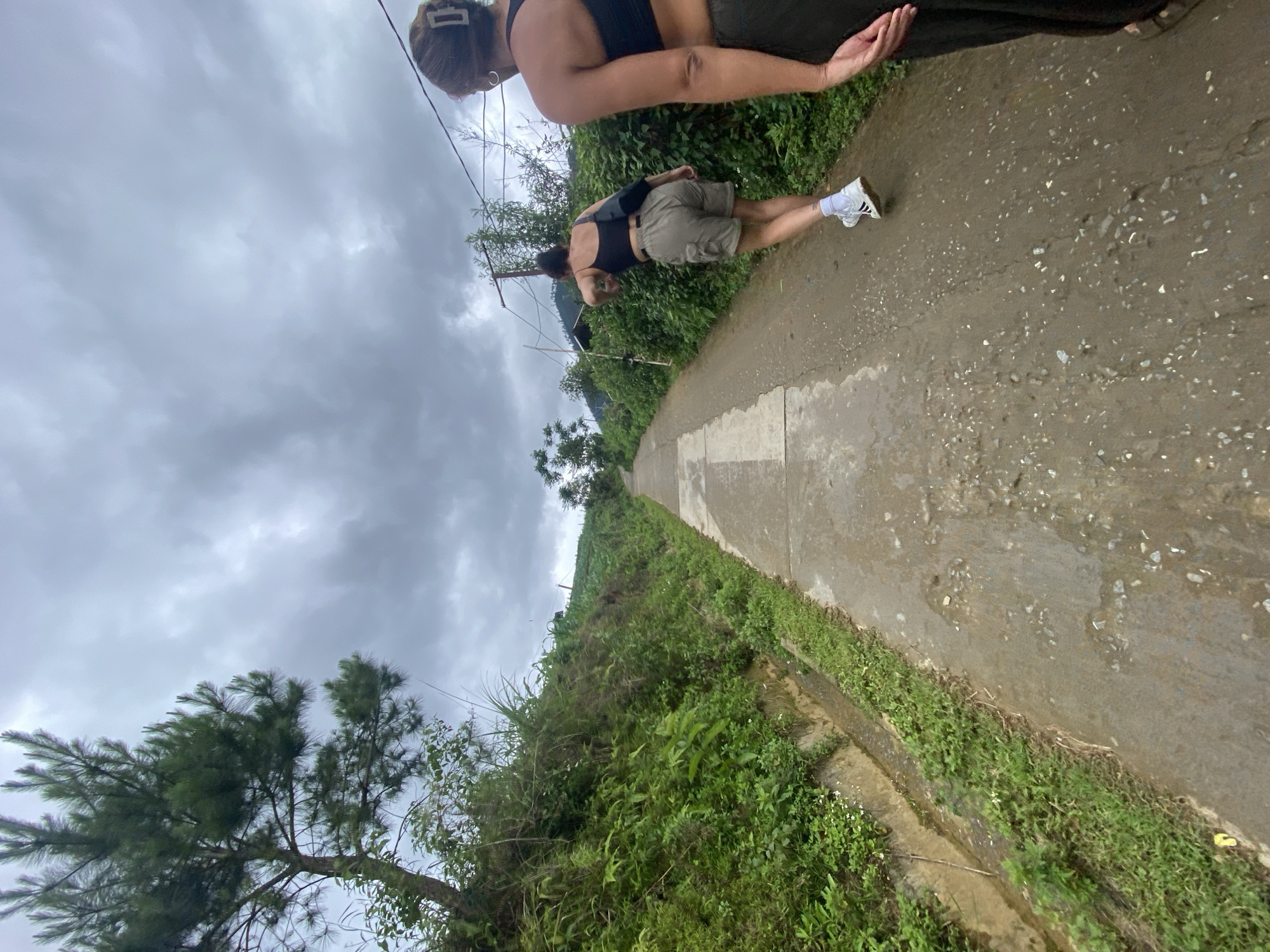 Two individuals walk along a concrete path in Dế Xu Phình, Vietnam, under a heavily overcast, grey sky. The path, visible from a low-angle perspective, has wet and muddy patches with small stones embedded. To the immediate left of the path is a narrow, earthen drainage ditch, beyond which dense green foliage and tall grasses grow on an upward sloping bank. A mature pine-like tree with dark green needles is prominent on the upper left. To the right of the path, a similar bank rises, covered in dense green vegetation, including what appears to be cultivated fields higher up the slope. Several rudimentary utility poles with wires strung between them are visible on this right side.
In the immediate foreground, facing away from the camera and positioned on the right side of the path, is a person whose back is visible from the shoulders down. This individual wears a dark-colored, possibly black, sports bra or crop top and dark lower garments that extend out of frame. A light-colored, rectangular hair clip is visible in their hair, which is styled upwards.
Slightly ahead and to the left of the foreground individual, a second person walks away from the camera. This individual wears a black spaghetti-strap crop top, light brown or khaki cargo shorts, white crew socks, and white sneakers with black accents. A black cross-body bag is slung over their left shoulder, resting on their lower back. Their head is turned slightly to the right, and their left arm is bent, reaching towards the green vegetation on the right bank. Both individuals are in motion, moving away from the camera's perspective.