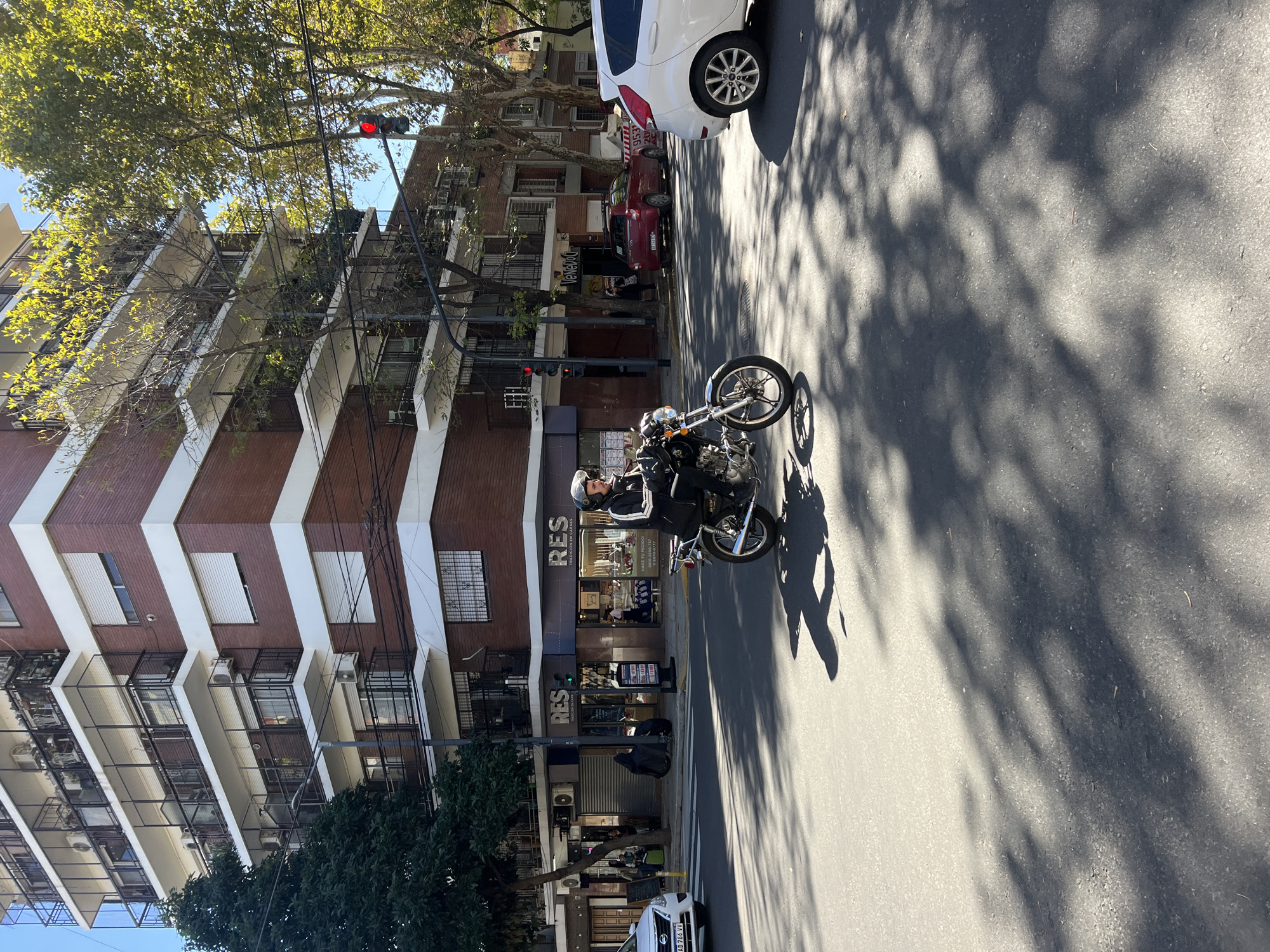 The image depicts a sunlit urban street scene in Buenos Aires, Argentina, featuring a mix of residential and commercial buildings. The perspective is from street level, looking down the road.
In the foreground and midground, a motorcyclist, wearing a helmet and dark jacket, is positioned towards the center-right of the frame, appearing to be stopped on a dark-colored motorcycle with chrome wheels. The street, made of asphalt, is heavily dappled with distinct shadows cast by overhead trees, indicating bright sunlight. The motorcyclist's shadow is also visible on the pavement.
To the right, a white car is parked, facing away from the viewer, with its rear visible. Its license plate, "AB 766 VV", is partially discernible. Further down the street, a red car is also parked.
The left side of the street is lined with multi-story buildings, characteristic of apartment complexes. They feature brick facades, white horizontal and diagonal trim, and multiple balconies, many of which are enclosed by metal grates. Air conditioning units are visible on several balconies. Ground-level storefronts are integrated into these buildings. One prominent storefront displays "RES TRADICIÓN EN CARNES" (RES Tradition in Meats) along with a phone number "0800-080-3337". Another visible street number is "1563". Overhead, green tree canopies obscure parts of the buildings and sky. Power lines are visible running across the scene. A traffic light, displaying a red signal, is positioned high above the street amidst the trees, suggesting an intersection.
The bright sunlight, strong shadows, and clear sky indicate a sunny day, likely morning or late afternoon.