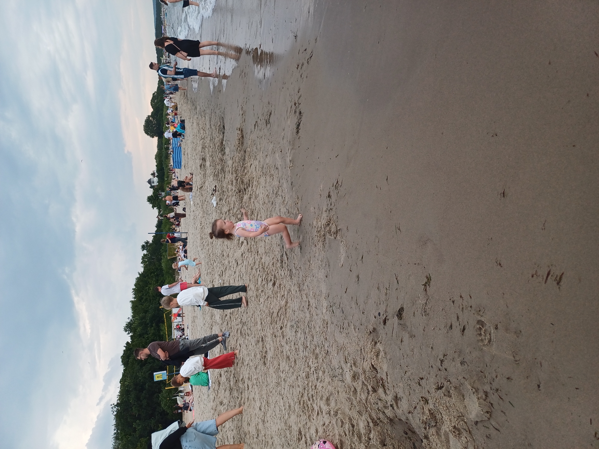 The image depicts a busy beach scene with numerous people enjoying the sand and water. In the foreground, a young girl in a swimsuit is walking on the wet sand near the water's edge. To the left, two young boys in white shirts and dark pants are standing on the sand. Further back, a man in a dark shirt and grey pants stands with his arms crossed, facing away from the camera. Other individuals and groups are scattered across the beach, some sitting, some walking, and some standing in the shallow water. In the background, trees line the edge of the beach, and the sky is overcast with clouds. This scene is likely at one of Gdańsk, Poland's popular beaches, such as Stogi Beach or Brzeźno Beach, given the typical summer atmosphere and the presence of beachgoers.