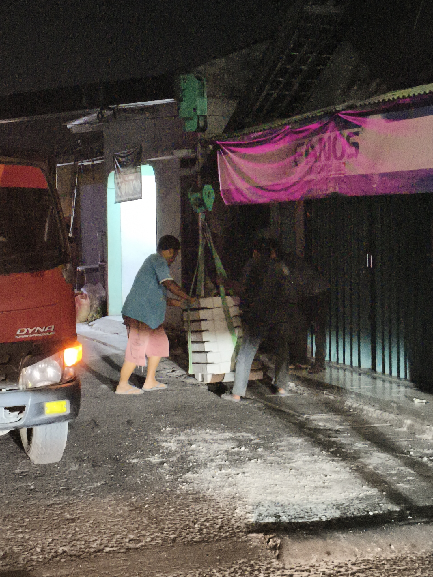In Depok, Indonesia, two individuals are engaged in material handling under artificial lighting. A red truck, identified as a "DYNA TURBO INTERCOOLER," is parked to the left of the frame, its headlights illuminated. A green electric hoist is suspended from an overhead beam, lifting a stack of pale rectangular objects. One person, wearing a blue shirt and pink shorts, is positioned to the right of the truck, appearing to guide the load as it is lifted. Another figure, largely obscured by shadow and motion blur, stands further to the right, also seemingly involved in the operation. The ground is covered in a dark, granular material with scattered white debris. A large pink banner with purple lettering is visible hanging from an overhead structure in the background. The scene suggests an outdoor industrial or commercial setting, possibly a construction site or warehouse loading area, at night.