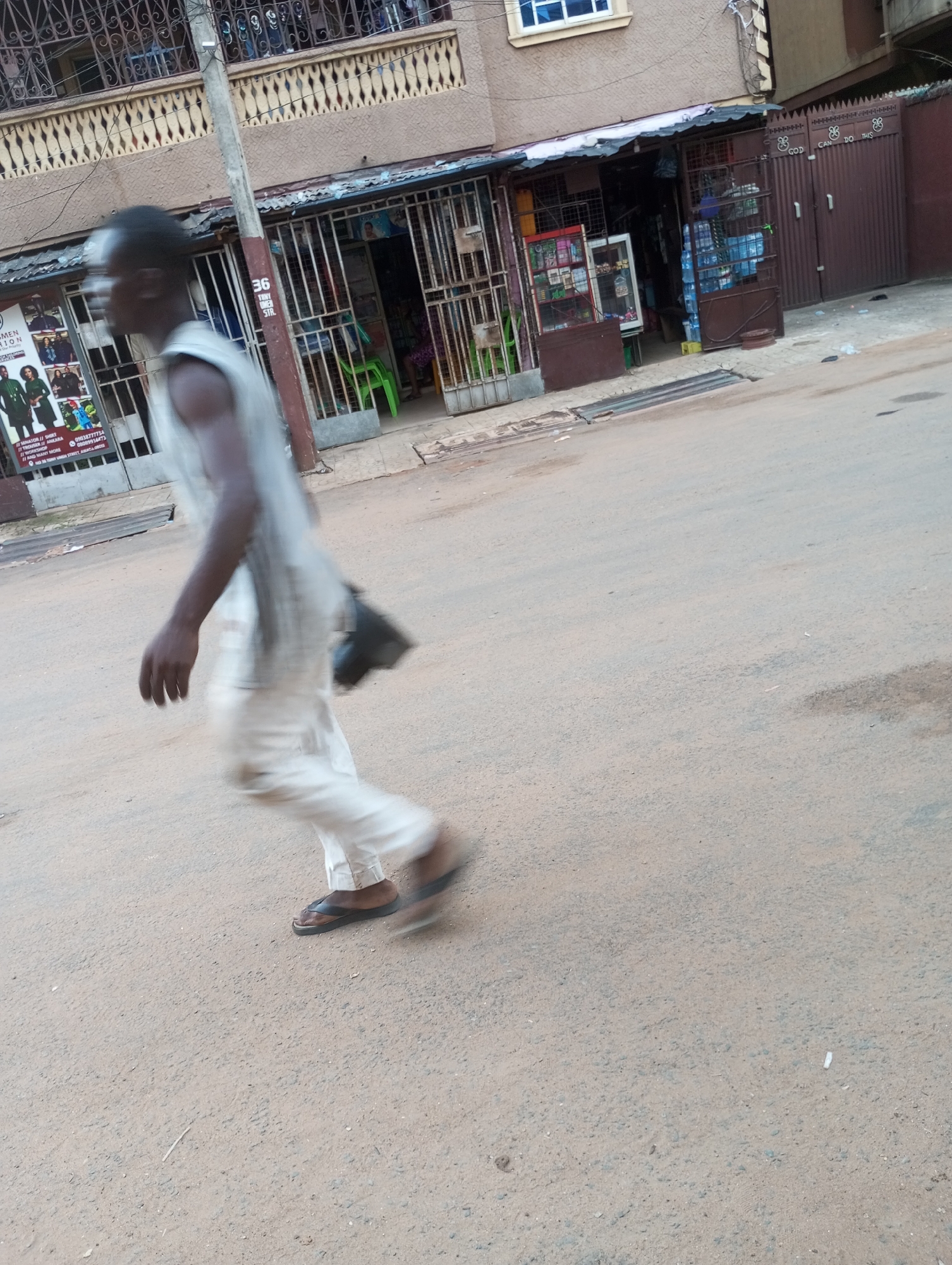 The image depicts a dynamic street scene in Onitsha, Nigeria, captured during what appears to be daytime. The primary subject is a dark-skinned male figure in motion, walking from the left to the right side of the frame. He is dressed in light-colored attire, consisting of a sleeveless top and trousers, and wears dark flip-flops. A small black bag or pouch is visible hanging at his side, and his movement is conveyed by a slight blur.
The background features a two-story building with a light-brown facade and decorative ironwork on the upper-level balconies. The ground floor houses several commercial establishments, secured by metal gates. On the left, a large sign is visible, advertising "WOMEN FASHION" and "MEN FASHION," along with other items like "TROUSER" and "SHIRT," under the banner "AND MANY MORE." Contact numbers "09038777734" and "08009914473" are also legible on this sign. A utility pole stands centrally on the left, bearing the number "36."
To the right, another storefront behind a metal gate displays goods, including stacks of bottled water. Adjacent to this is a large, dark brown metal gate that clearly reads "GOD CAN DO THIS." The street surface in the foreground and midground is a light-colored asphalt or packed dirt, showing some wear and tear. The overall setting suggests a busy, urban environment with local businesses.