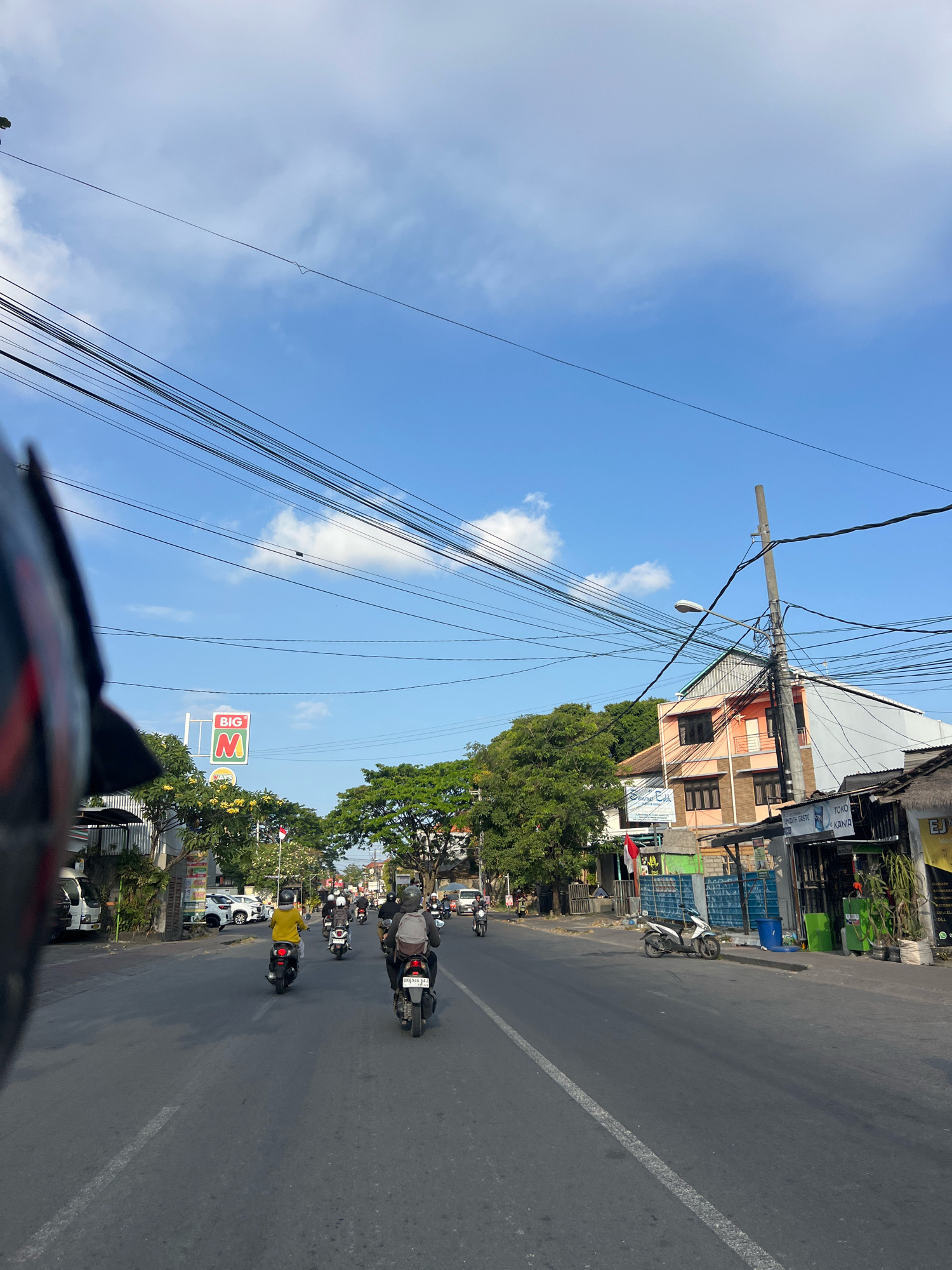 The image depicts a street scene in Denpasar Barat, Indonesia, under a clear blue sky with scattered clouds. The perspective is from a person riding a motorcycle, indicated by the partial view of a black motorcycle helmet in the foreground. The street is paved with asphalt and has a white dashed line marking the lanes.
Several people are riding motorcycles along the street, with most wearing helmets. Some of the motorcyclists are individuals, while others appear to be riding in groups. One person in the middle of the road is wearing a bright yellow shirt and a helmet. Another motorcyclist in the foreground, also wearing a helmet, has a backpack on.
Along the sides of the street, there are buildings and trees. A prominent sign with "BIG M" in red and green letters is visible. There are also shops and businesses, some with awnings or overhangs. One building is a multi-story structure with a peach-colored facade. Utility poles with numerous wires crisscross overhead, a common sight in urban areas. Various parked vehicles, including cars and motorcycles, are visible on the side of the road. A Indonesian flag is also displayed near one of the buildings. The overall impression is a busy, everyday street in an Indonesian city.