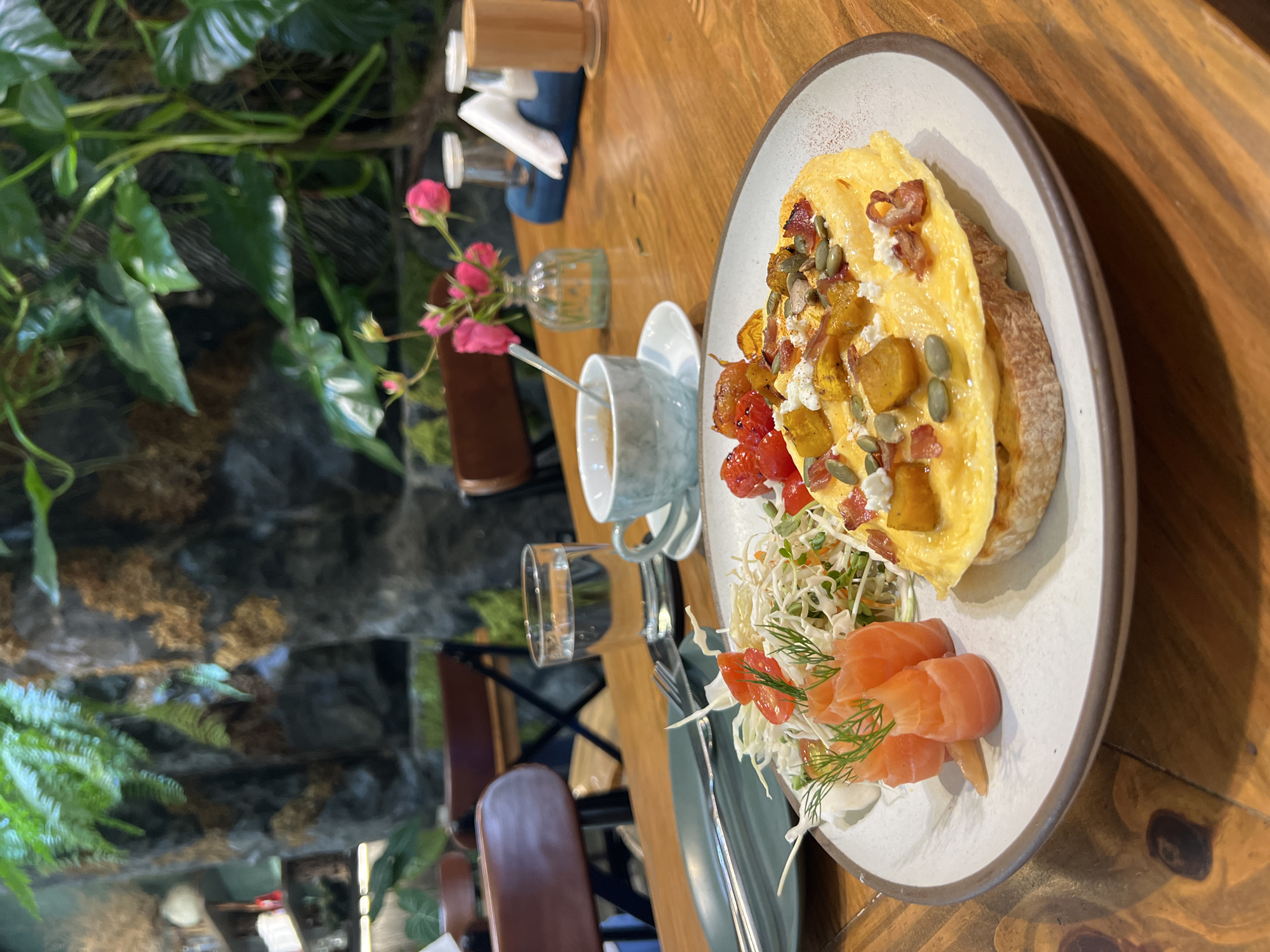 The image depicts a partially eaten breakfast dish served on a ceramic plate atop a wooden table. The breakfast dish consists of an omelet served on bread with butternut squash, pumpkin seeds, goat cheese, and bacon on top. A portion of sliced salmon with sprouts and dill is also present on the plate. Nearby are a glass of water, a cup of coffee or tea with a spoon, a small vase of pink flowers, a dispenser of what appears to be paper napkins, and silverware. The setting appears to be a restaurant or cafe in Nha Trang, Vietnam, judging by the foliage and stone wall visible in the background. The image has a blurred, slightly tilted perspective and the setting appears to be inside. No people are in the shot.