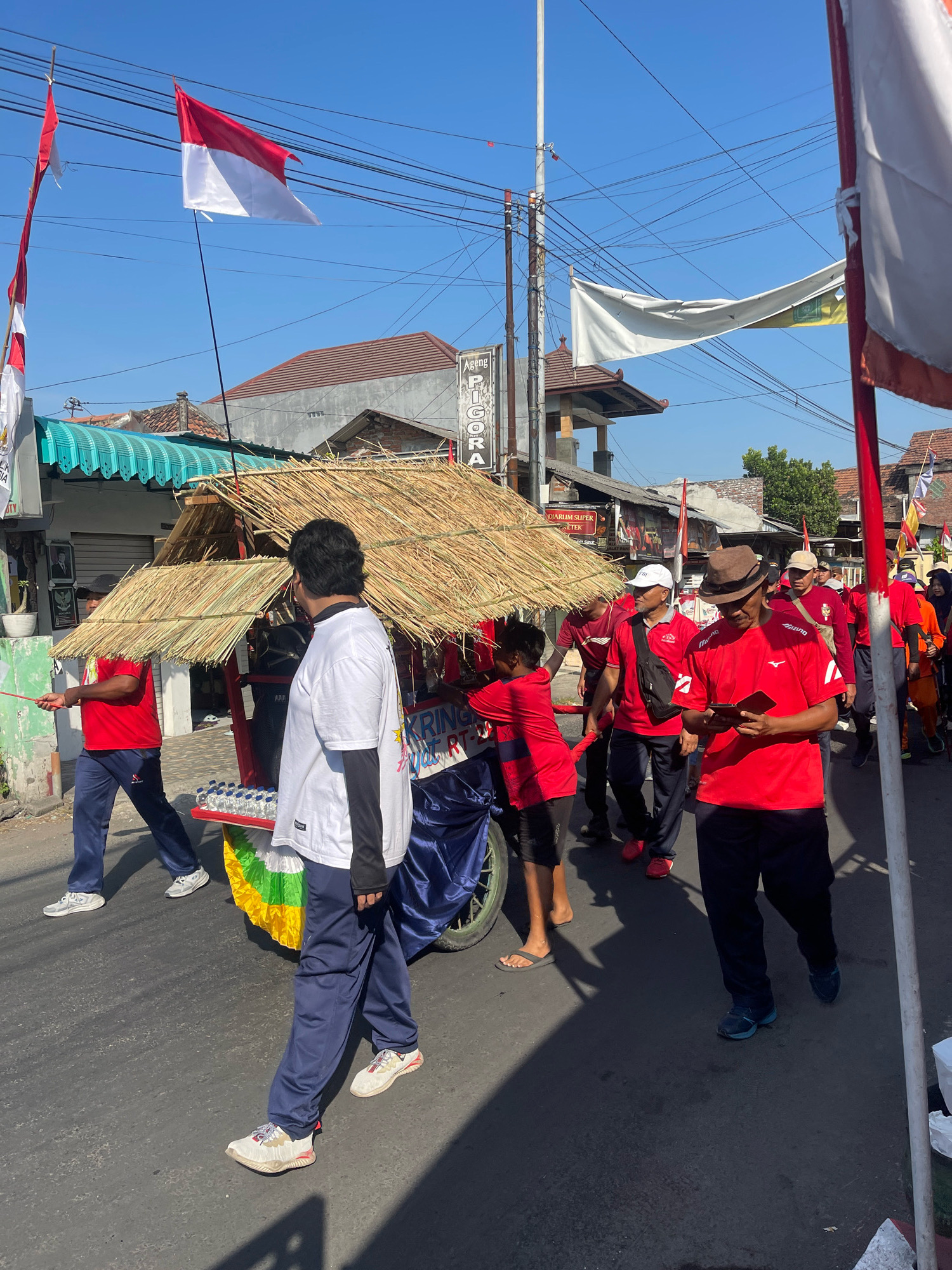 In the city of Candi, Indonesia, a procession is underway on a paved street under a clear blue sky. The foreground is dominated by a group of people walking, some of whom are wearing red shirts and dark pants. One individual in the front is wearing a white t-shirt with blue sweatpants. Behind them, a cart with a thatched roof, possibly a mobile refreshment stand, is being pushed. The cart has a display of bottled water. Several Indonesian flags are visible, both on poles and as part of banners. Buildings line the street, with one having a green-tiled roof and another appearing to be a shop with signage. The overall scene suggests a community event or celebration.