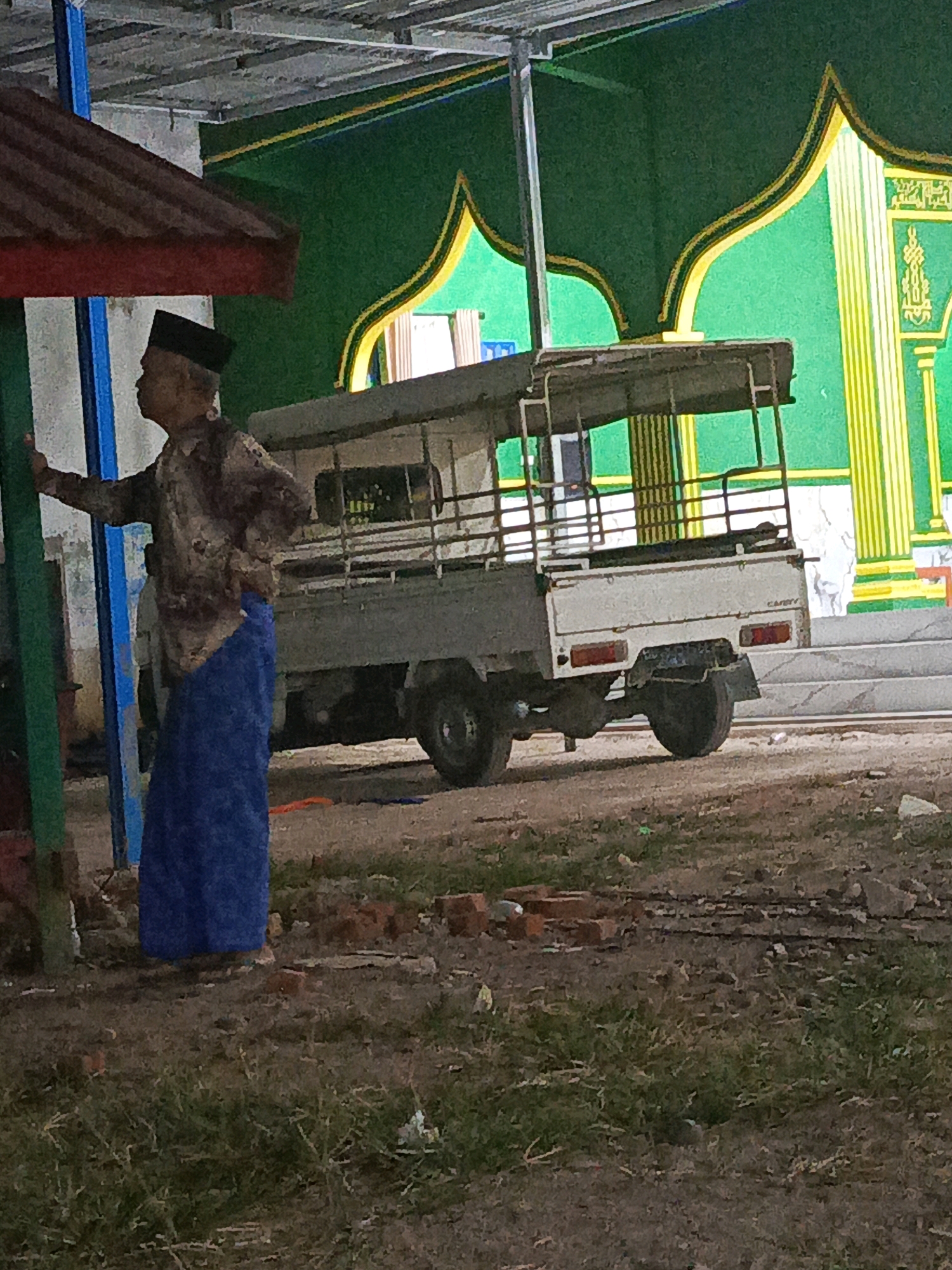 An Indonesian man wearing a black peci (cap) and a brown patterned shirt over a long blue sarong stands to the left of a white pickup truck with a metal cage. The man is facing left and appears to be resting his left hand on a green pole. The truck is positioned behind him and to his right. The background features a green wall with yellow and white decorative elements resembling arches and pillars, consistent with the architectural style of a mosque. The ground is rough and covered with dirt, grass, and some scattered bricks. The overall scene appears to be outdoors, possibly in a courtyard or parking area adjacent to a religious building in Megang Sakti V, Indonesia.