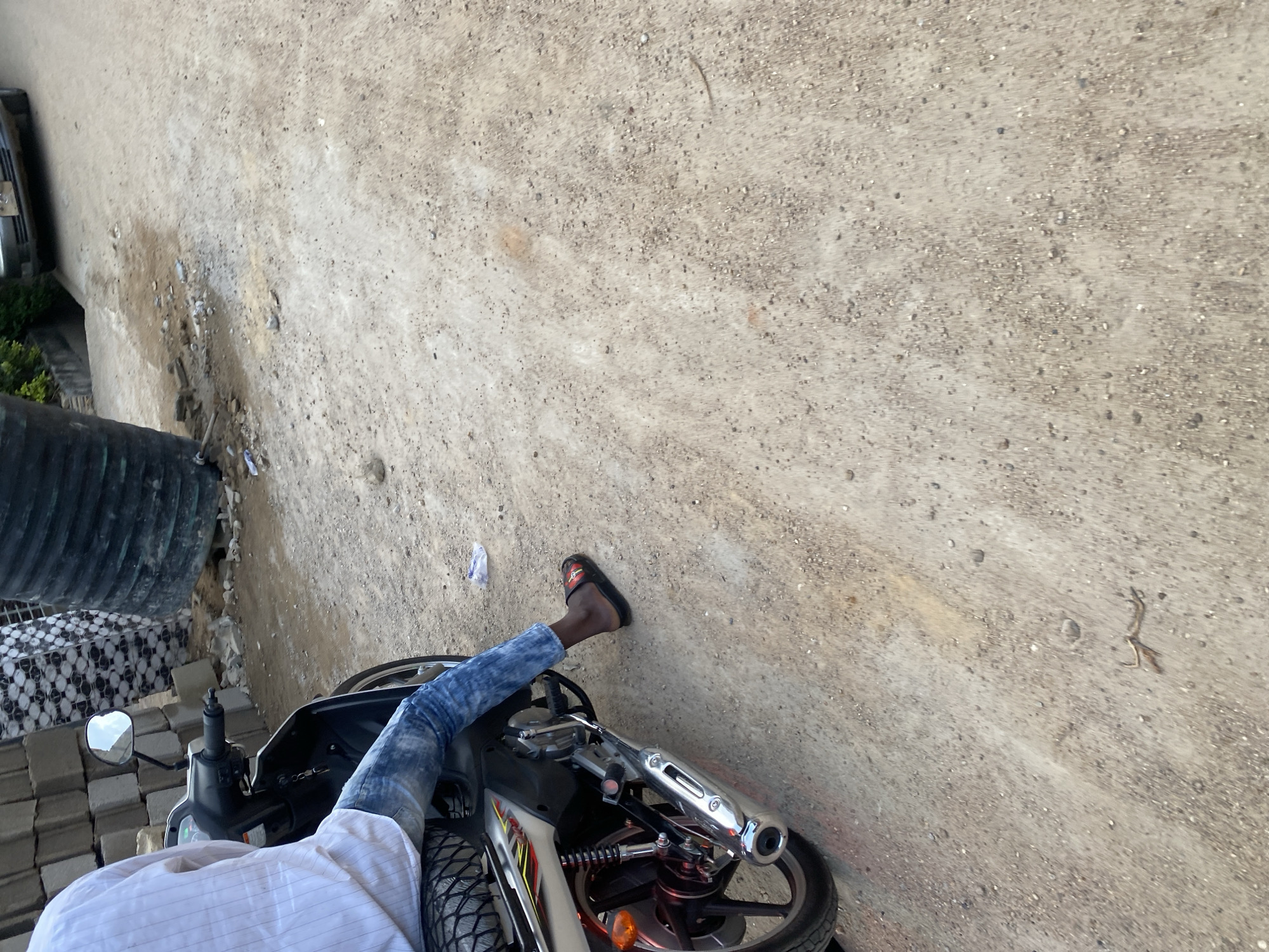 A person is sitting on a motorcycle on a dirt road in Jos, Nigeria. The person is wearing a white striped shirt and blue jeans. The motorcycle is silver and black, with a chrome exhaust pipe. In the background, there are stacks of gray bricks and a large black water tank. A car is visible further down the road. The ground is covered in gravel and dirt.