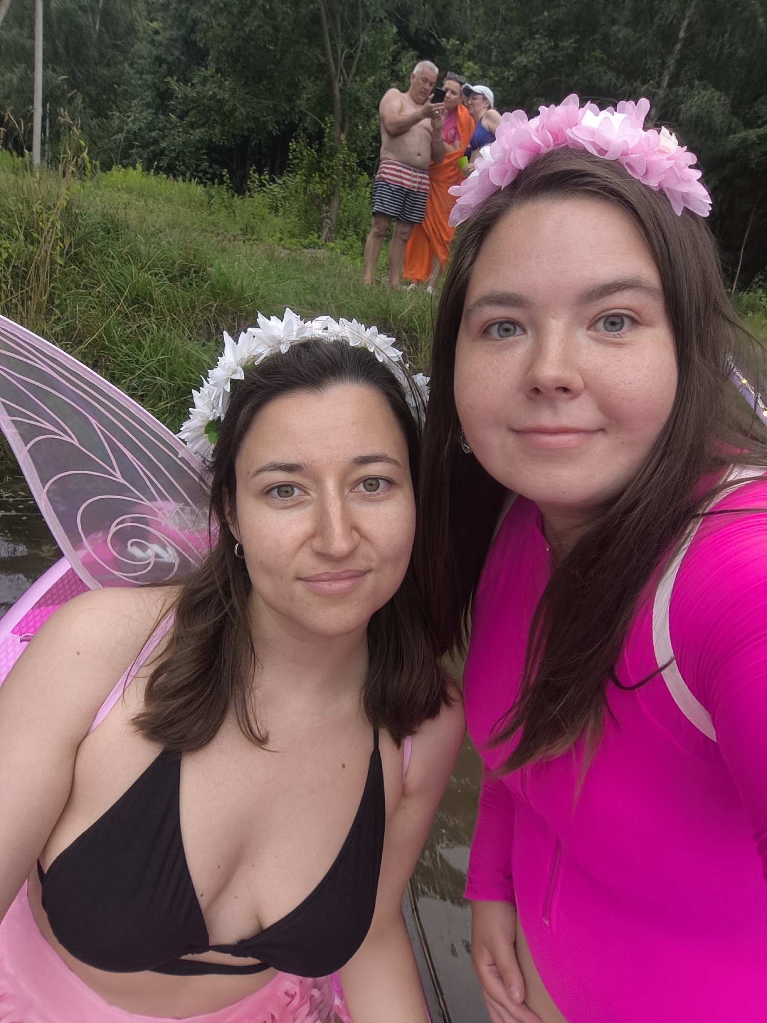 Two women, appearing to be in their late teens or early twenties, are posing for a selfie in an outdoor, grassy environment with a body of water in the foreground. Both women are wearing flower crowns, and one has fairy wings attached to her back. The woman on the right is wearing a bright pink long-sleeved outfit. The woman on the left is wearing a black bikini top and a pink skirt. In the background, further away, three other individuals are visible. One man, shirtless and wearing striped shorts, is holding up a phone, seemingly taking a picture. Next to him, two other people, one in an orange garment and another in a blue top and white hat, are also present. The overall setting suggests a casual outdoor gathering, possibly a themed event. The location is not identifiable as Kaliningrad, Russia.