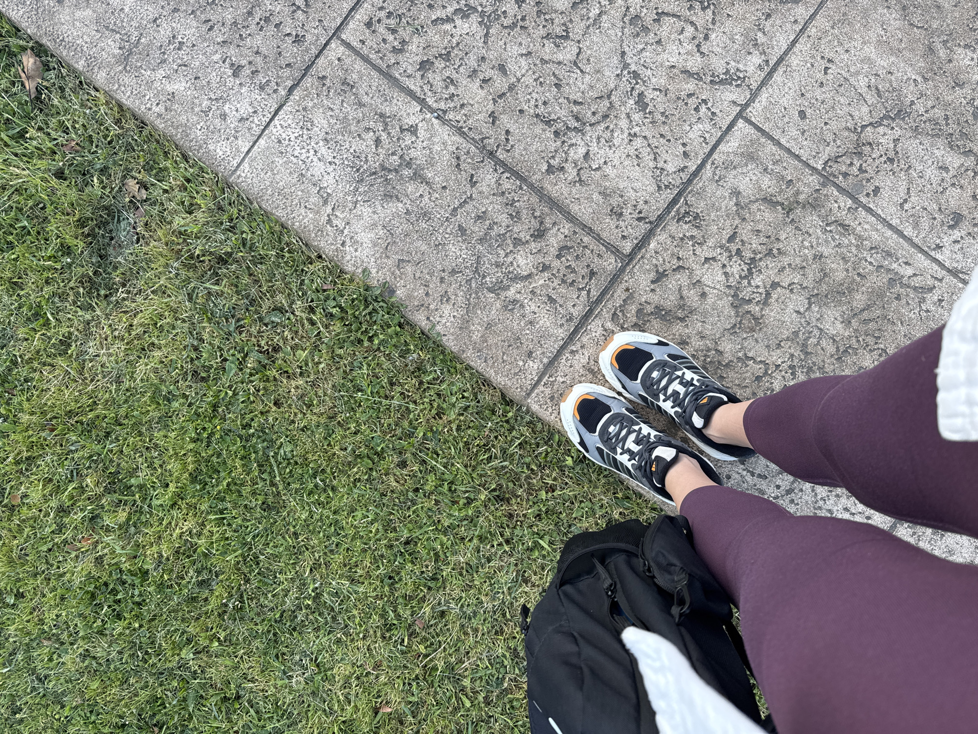 A person stands at the edge of a grey stone-tiled pathway, where it meets a grassy area. The person wears maroon leggings and black, grey, and white Adidas sneakers with a tan accent on the sole. A black backpack rests against the person's right leg, partially obscuring the leggings. The grass is green and appears neatly trimmed. The stone pathway is textured, and the image is taken from an overhead perspective.