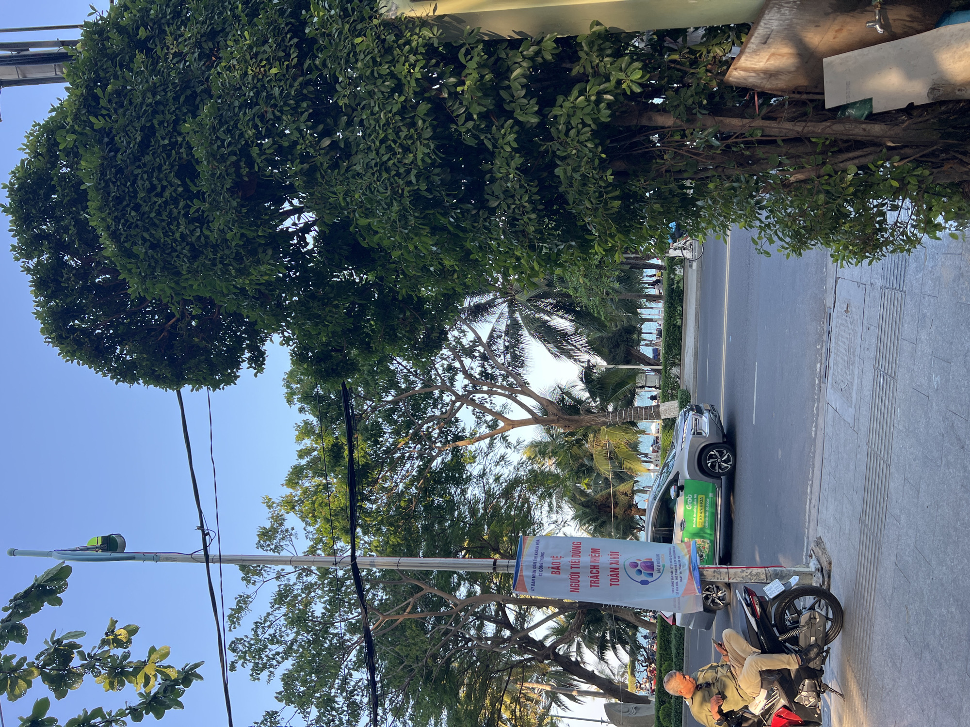 The image depicts a street scene in Nha Trang, Vietnam, with a road running alongside the beach. A silver car with green Grab advertising is parked on the side of the road next to a scooter on which a man is sitting. He appears to be using his phone. A banner ad is attached to a utility pole, bearing the text "ỦY BAN NHÂN DÂN TỈNH KHÁNH HÒA" and the words "NGƯỜI TIÊU DÙNG / TRÁCH NHIỆM / TOÀN XÃ HỘI". Palm trees and other greenery are visible in the background, near the beach. The sky is clear and blue.