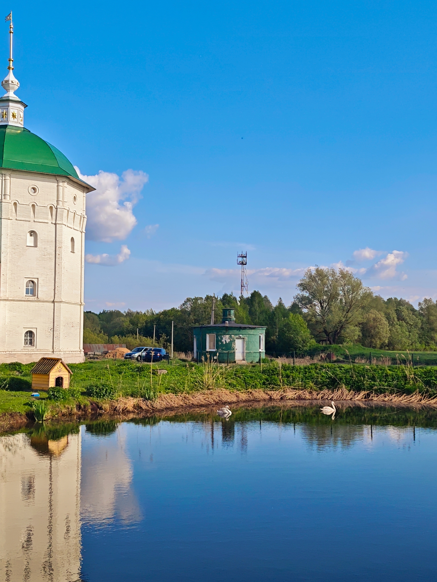 The image depicts a scene in Луговой, Russia, featuring a white brick tower with a green dome, reflecting in a body of water. To the left of the tower is a small yellow dog house. In the background, there's a green outbuilding, cars, and trees. An antenna tower rises from the trees. Two white swans are visible on the water's surface. The sky is blue with some clouds.