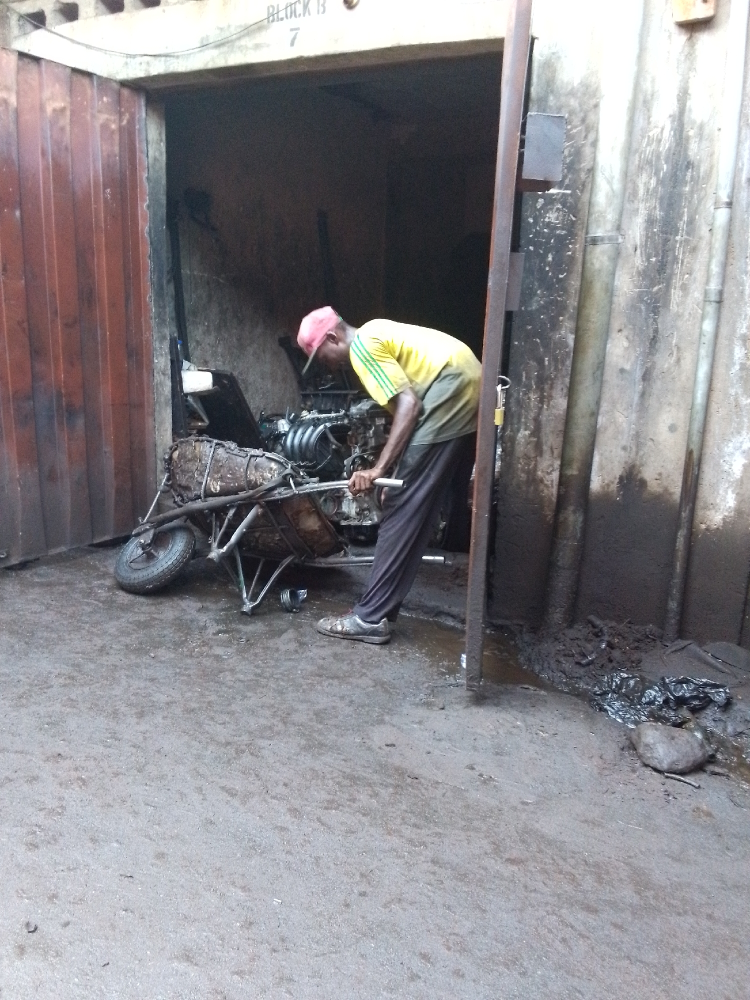 This media file captures a man working in what appears to be a mechanics workshop or repair area in Onitsha, Nigeria. The scene is set outdoors, partially under the cover of a building structure.
**Subjects:** The main subject is a dark-skinned man, seen from a side-back angle, leaning over a disassembled engine. He wears a yellow and green short-sleeved shirt, dark trousers, a pink cap, and dark shoes.
**Objects:** A heavily used, metal wheelbarrow holds a significant portion of an engine, including its manifold and other mechanical parts. The wheelbarrow itself looks old and grimy. Other objects include the corrugated metal doors of the workshop, concrete walls, metal support beams, and various debris on the ground, such as a black plastic bag and a large stone.
**Scene and Setting:** The setting is an industrial or workshop environment. The ground is concrete, appearing damp and dirty in places. The walls of the building are discolored and show signs of wear and grime. The workshop entrance has a large, open corrugated metal door.
**Activity:** The man is actively engaged in working on the engine. He is bent over the engine, holding onto a metal component, suggesting he is either examining, repairing, or assembling it.
**Notable Details:** The lighting indicates it is daytime, although the interior of the workshop is relatively dim. The overall impression is of a busy, practical workspace. The ground on the right side of the image is particularly muddy and littered.
**Visible Text:** Above the entrance to the workshop, on a concrete beam, the text "BLOCK B 7" is visible.