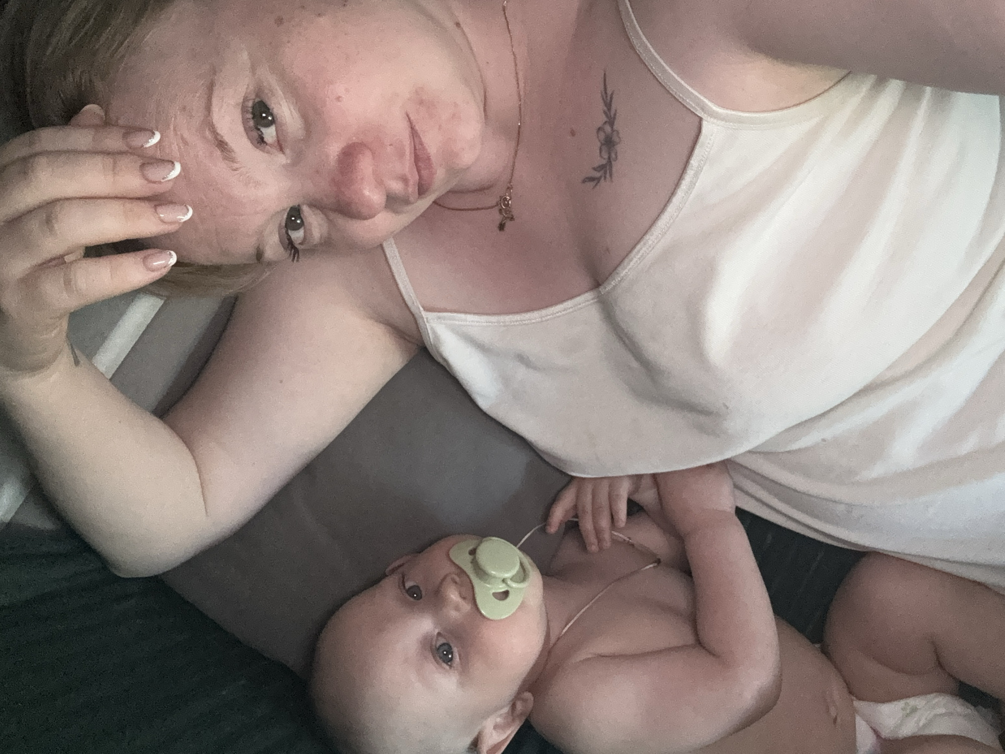 A woman and a baby are lying on what appears to be a bed or soft surface. The woman is fair-skinned with blonde hair. She has a flower tattoo on her chest, and she wears a delicate gold necklace. The baby has a pacifier in its mouth and a thin cord around its neck, possibly attached to the pacifier. The background shows a dark surface. There is no indication that the image was taken in Chaikovsky, Russia, or any city at all.