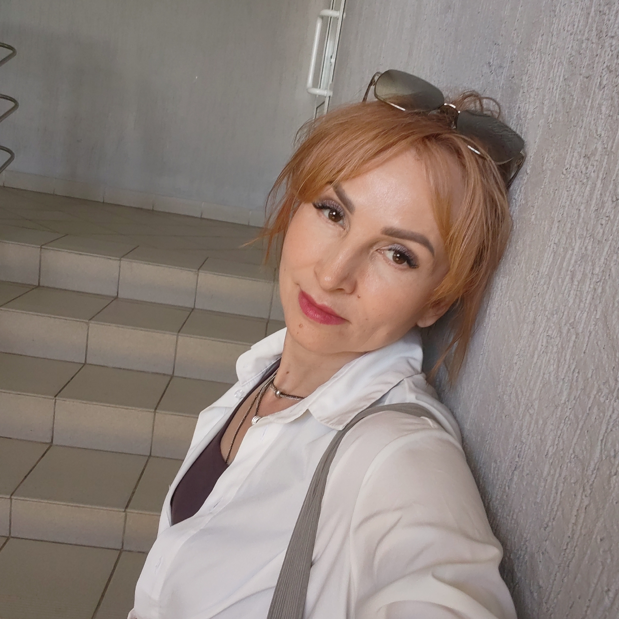 A woman with auburn hair and sunglasses perched on her head leans against a textured, light-gray wall. She's wearing a white, collared shirt over a dark top and a gray strap crosses her shoulder. A staircase with light-colored tiled steps is visible in the background.
The location is Chaikovsky, Russia.