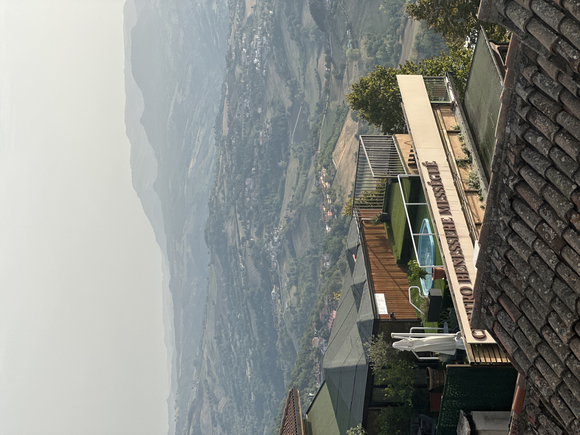 This is a photograph of the City of San Marino, San Marino, taken from a slightly elevated position looking down over a valley. In the foreground, there are several tiled rooftops, with one clearly visible that is grey and textured with lichen. Above these rooftops, there is a building with a light-colored facade bearing the text "CENTRO BENESSERE MESSÉGUÉ" in large, dark letters. This building has a rooftop area that includes a small blue swimming pool, artificial turf, and potted plants. Next to the pool, there is a white umbrella draped over a stand. Further back, the landscape unfolds into rolling hills covered in green vegetation and dotted with small villages. In the far distance, hazy mountains complete the panorama under a pale, overcast sky. The overall impression is one of a tranquil, scenic vista.