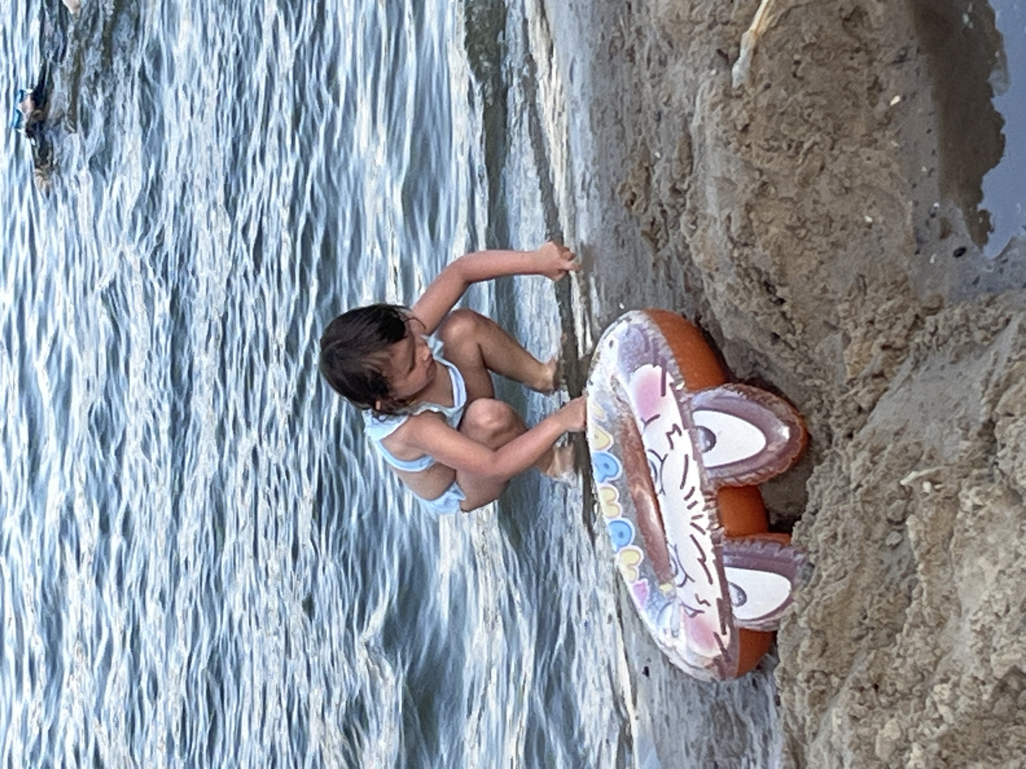 A young child is playing on a sandy shore next to a body of water. The child is wearing a light-colored swimsuit and is crouched down, reaching out a hand towards the sand. Beside the child is an orange inflatable ring with a cartoonish design, featuring large eyes and a smile. The water is rippling, suggesting a gentle current or waves. The sand appears damp and textured, with some small pebbles or debris visible. In the background, a person can be seen partially submerged in the water. The lighting suggests it is daytime, possibly late afternoon due to the soft light. The scene evokes a sense of carefree play and summer enjoyment. The overall location appears to be a natural beach or riverbank.