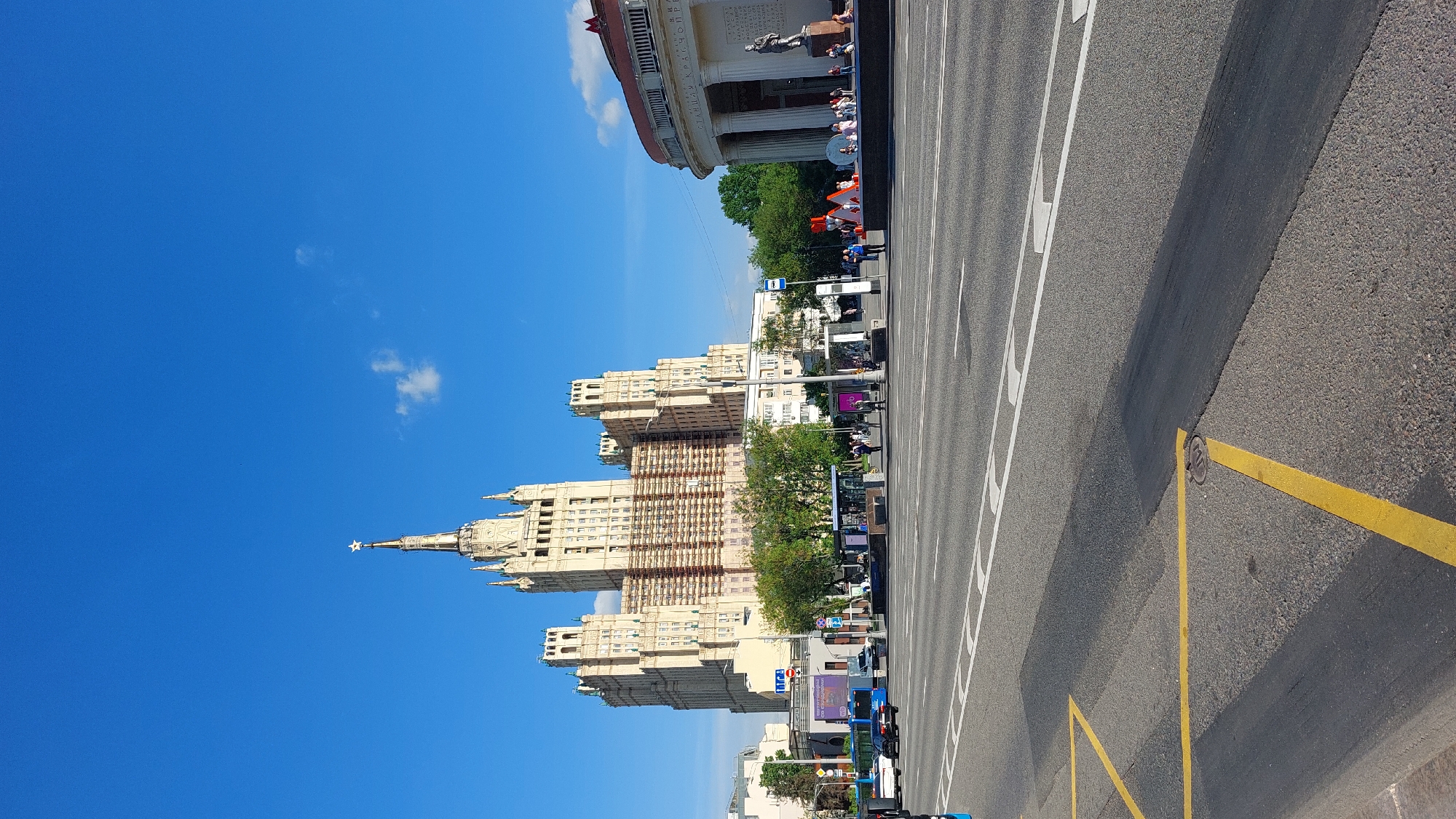 The image shows a view of Kotelnicheskaya Embankment Building in Moscow, Russia. The building, one of the Seven Sisters skyscrapers, dominates the left side of the frame against a clear blue sky. Its exterior appears partially under renovation with scaffolding visible. To the right, a wide road with multiple lanes is marked with white and yellow lines. Pedestrians are present on the sidewalks alongside trees and other buildings. In the background is a building with columns.