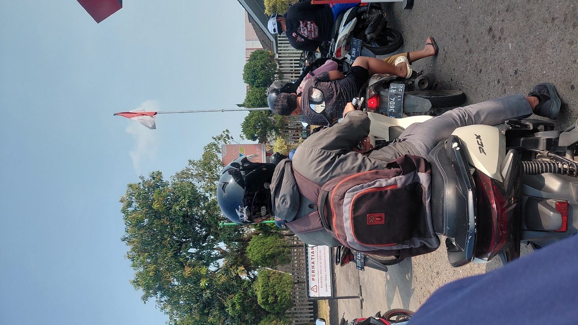 In Rejasari, Indonesia, under a clear blue sky, a group of people on motorcycles are stopped in traffic. The foreground is dominated by the back of a rider wearing a black helmet and a grey jacket over a backpack. The motorcycle is white with "PCX" branding. Behind this rider, another motorcycle carries two passengers, one of whom is a child wearing a patterned t-shirt. Further back, a third motorcycle has a rider wearing a black t-shirt with Indonesian text and a white cap. A red and white Indonesian flag is visible flying on a flagpole in the background, amidst trees and buildings. A warning sign with text in Indonesian is also present. The scene depicts a common urban traffic situation with multiple motorcycles.